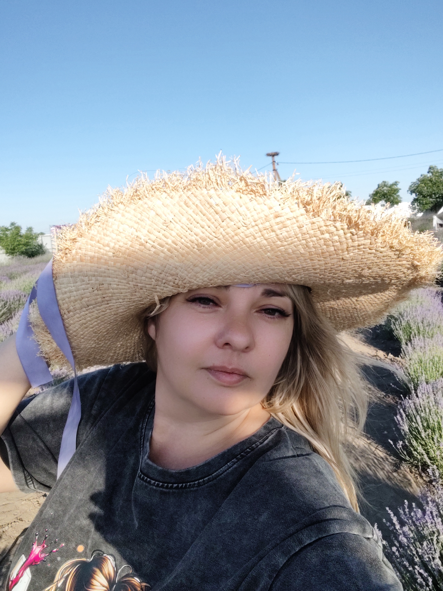 In this selfie taken outdoors, a woman with blonde hair is captured from the chest up. She is wearing a large, woven straw hat with a frayed edge and a light purple ribbon. The hat casts a shadow over her face, obscuring some of her features, but her eyes and mouth are visible. She has makeup on, including visible eyeliner and mascara. Her expression is neutral. She is also wearing a dark gray t-shirt with a graphic print on the front. The background consists of rows of purple lavender plants, suggesting the location is a lavender field. The sky above is a clear, bright blue, indicating it is daytime, likely sunny. The sunlight creates shadows on her shirt and the ground. In the distance, there are some trees and buildings, and a utility pole is visible in the far background. The overall impression is a relaxed, outdoor scene, possibly during the summer months given the flowering lavender and clear sky. The location is identified as Nechaiane, Ukraine.