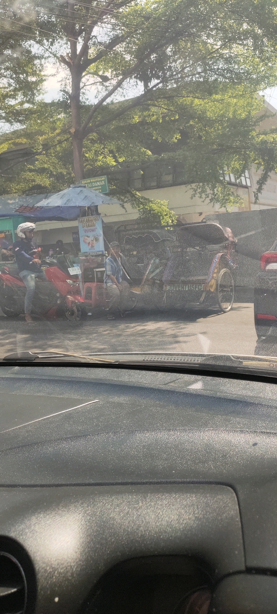 The image shows a street scene in Semarang, Indonesia, likely near a historical site given the signage that says "MAKAM WALIYULLAH." In the foreground, the dashboard and windshield of a vehicle are visible, suggesting the photo was taken from inside another car.
The middle ground features a street with several elements. A red motorcycle is parked, with a person wearing a white helmet and jeans sitting on it. To the right of the motorcycle is a small stall with a blue umbrella, selling drinks. A sign on the stall reads "ES JENDOL BU DWI." Behind the stall, a pedicab (known as a becak in Indonesia) is parked, with a person sitting on it. The pedicab has intricate decorations and text on its side that appears to say "JUVENTUS FC". A glimpse of another vehicle, possibly a red car, is visible to the far right.
The background is dominated by lush green trees, suggesting a warm climate. Behind the trees, a building with windows can be seen. The overall impression is a typical street view in a populated area of Semarang, possibly a tourist spot given the presence of the becak and the signage. The lighting suggests it is daytime, likely with bright sunlight filtering through the trees.