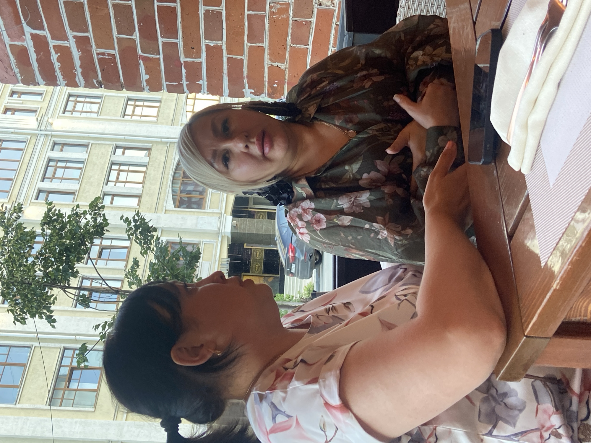 Two women are seated at a table outdoors, with a brick wall to their right and a building with multiple windows in the background. The woman on the left, with dark hair pulled back in a ponytail, is wearing a floral-patterned blouse and looking towards the woman on the right. The woman on the right, with blonde hair and dark earrings, is wearing a darker floral-patterned blouse and looking at the camera. A cell phone and a folded napkin with silverware are on the table in front of them. The overall scene suggests a casual dining or meeting setting.