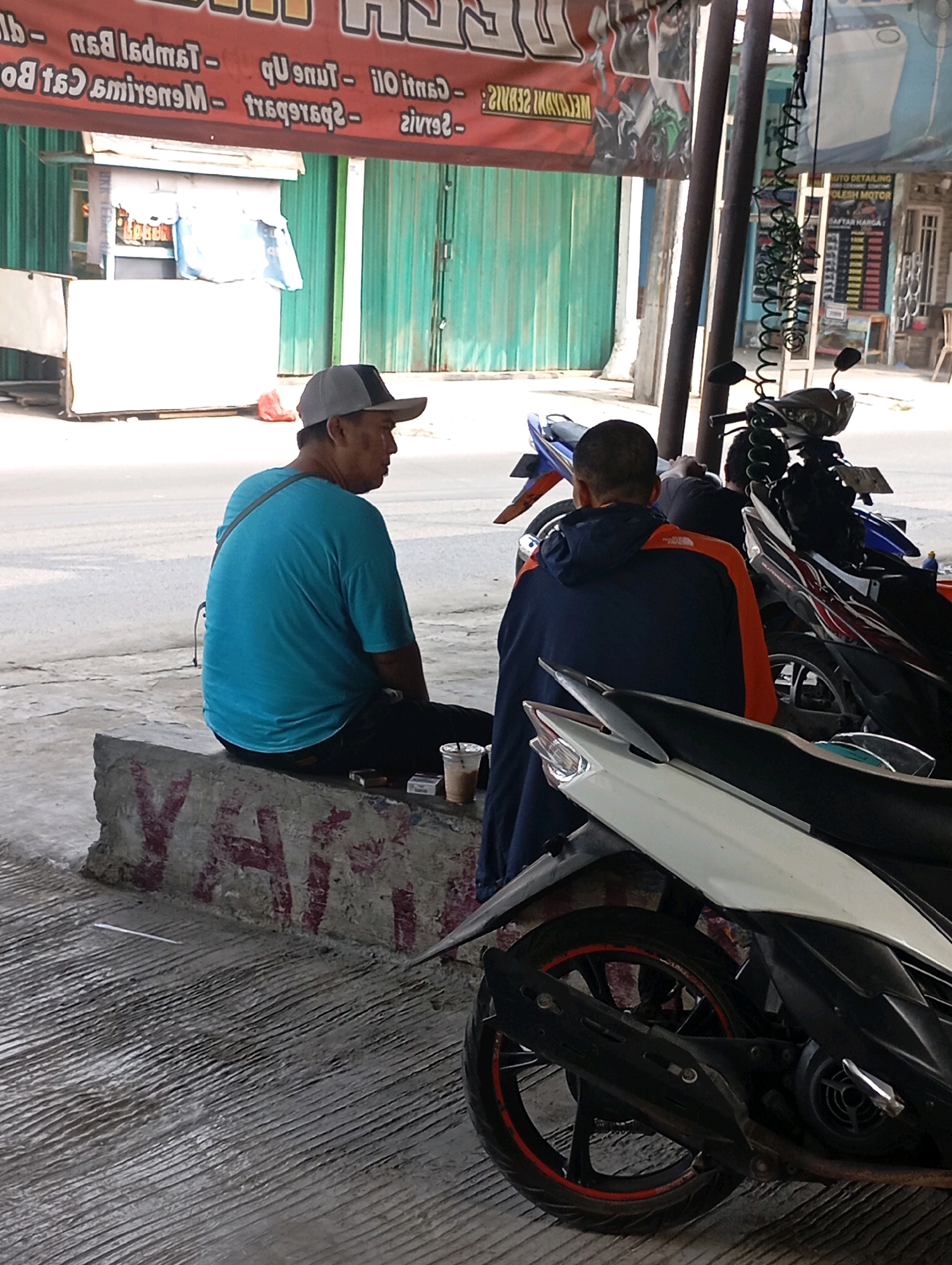 In Babelan, Indonesia, two men are seated outdoors next to motorcycles. One man, wearing a blue t-shirt and a grey cap, sits on a concrete barrier. Next to him on the barrier are a pack of cigarettes, a lighter, and a plastic cup with a beverage. The second man, in a blue and orange jacket, is seated on a motorcycle. The scene appears to be in an urban or commercial area, with a building and signage visible in the background. The ground is paved with concrete.