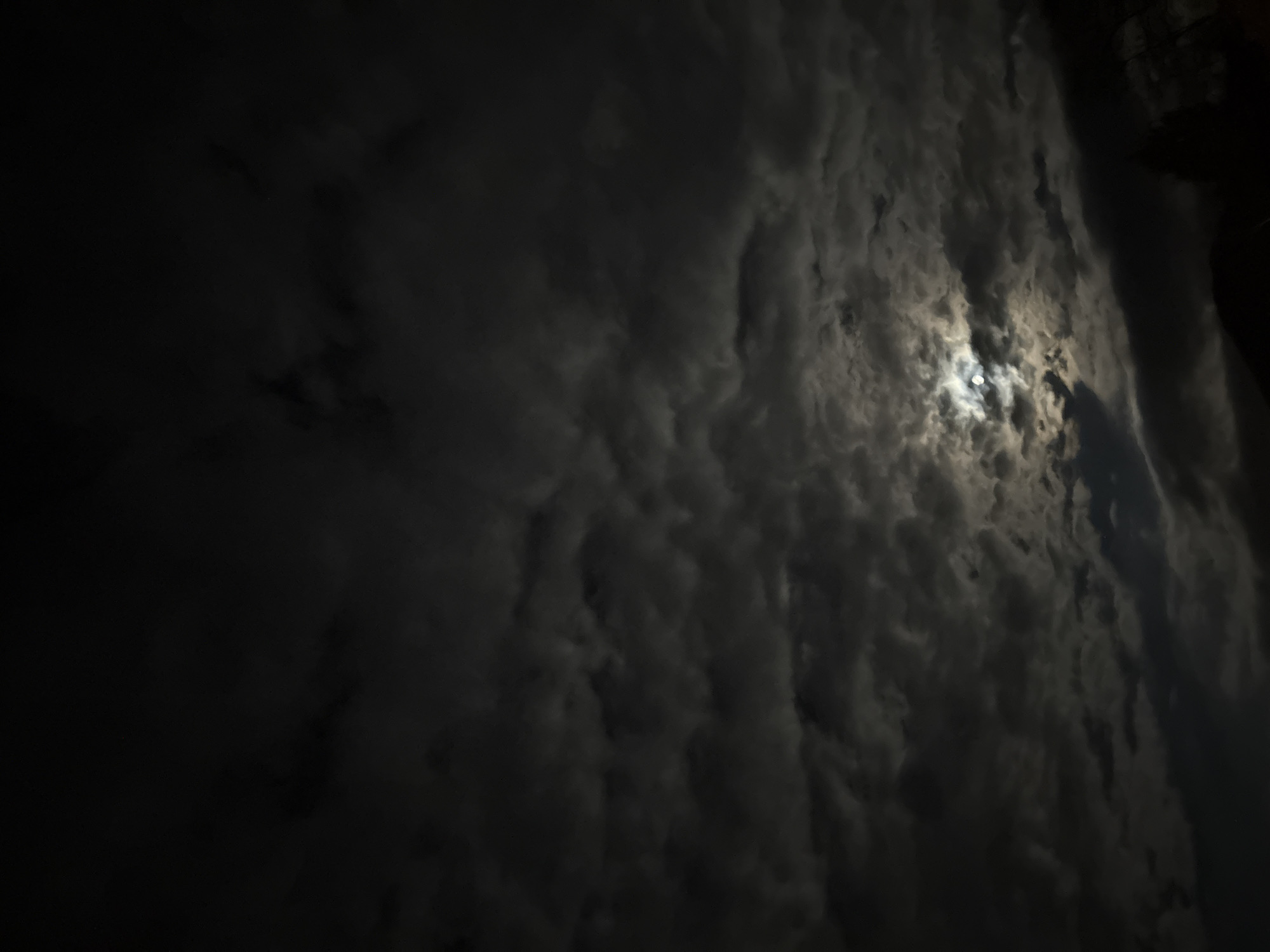The image depicts a night sky with a full moon partially obscured by dark, heavy clouds. A small portion of a structure is visible in the top right corner. The location appears to be Vrachesh, Bulgaria. No people are visible in the frame.