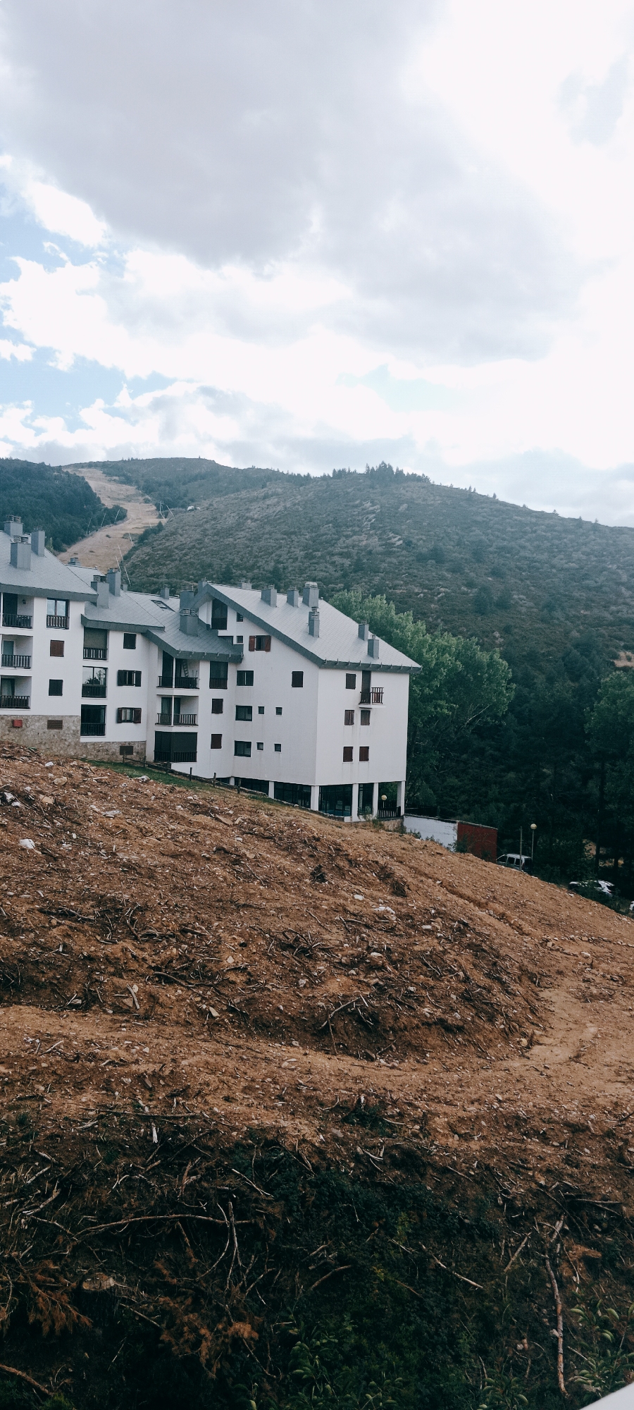 The image depicts a mountainous landscape with a modern white apartment complex in the foreground. The building has multiple stories, balconies, and numerous windows. The ground in front of the building is disturbed earth, with exposed roots and debris, suggesting recent construction or land clearing. In the background, a steep, tree-covered mountain rises, with a cleared path or ski slope visible on its upper slopes. The sky is partly cloudy, with patches of blue sky and white clouds. The location is identified as La Pinilla, Spain, a ski resort town known for its mountainous terrain and winter sports. No people are visible in the image.