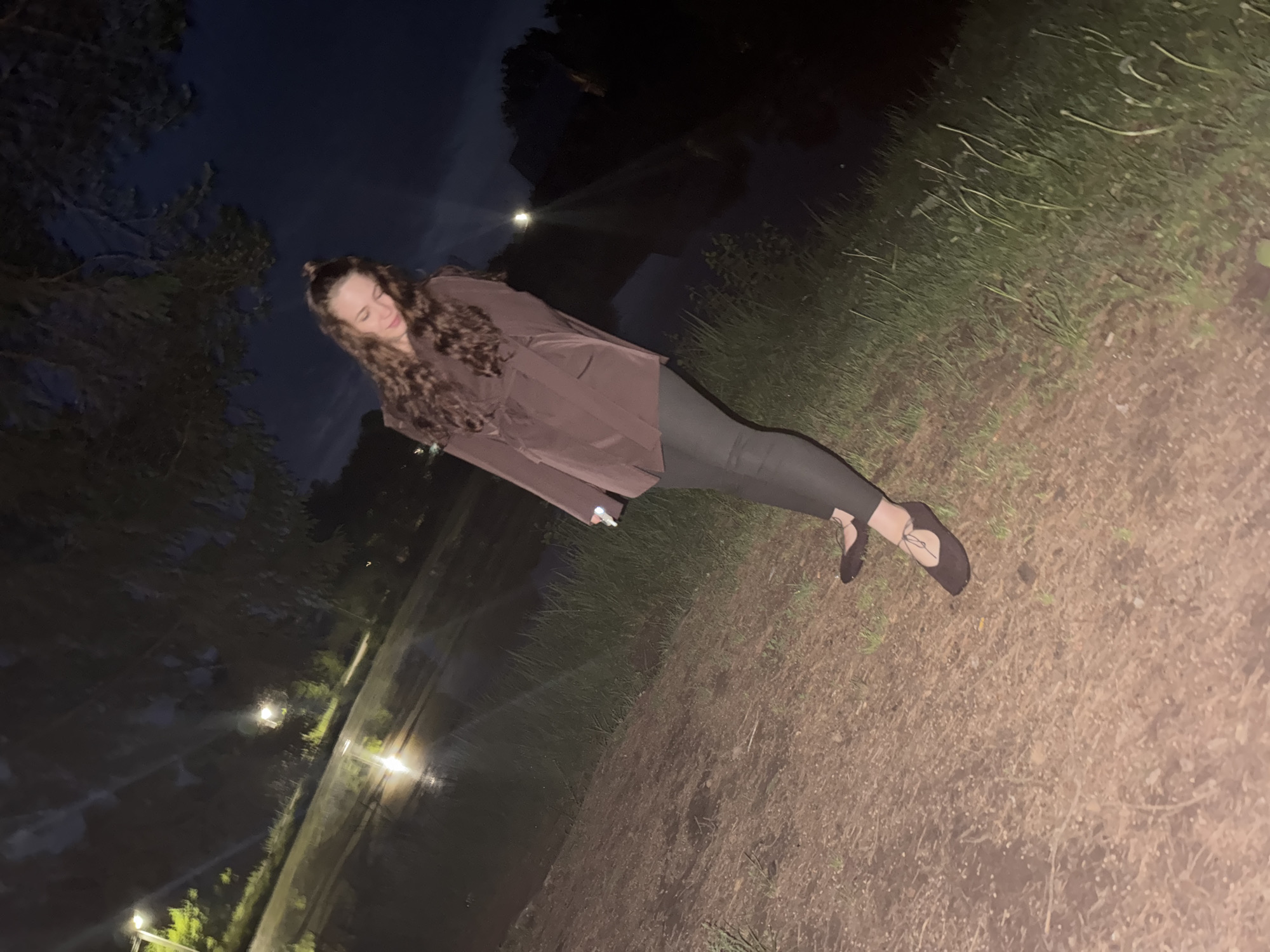 A woman with curly brown hair stands at an angle in an outdoor environment. She is wearing a brown blazer, black leggings, and black shoes. The background suggests a park or grassy area, with some visible trees and possibly a body of water with reflections from streetlights. The scene appears to be at night or in low light conditions.
Specific location information (Chaikovsky, Russia) is not possible to confirm from the image alone.