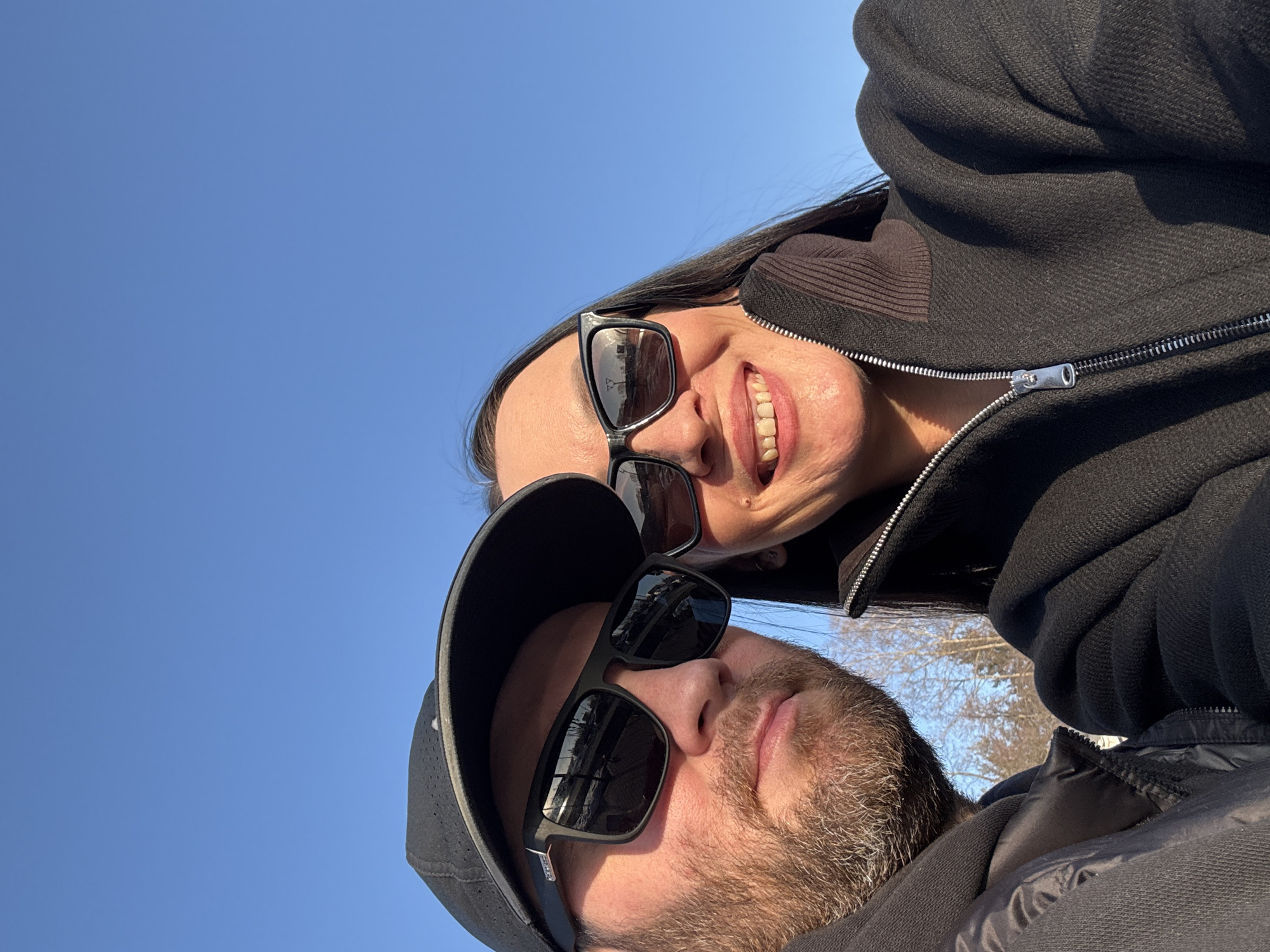 An outdoor selfie taken in the city of Chaikovsky, Russia, features two individuals, a male and a female, under a clear blue sky. The scene is illuminated by direct sunlight.
The male, positioned on the left, is visible from the chest up. He has a light brown and grey beard, wears a black baseball cap, and black framed sunglasses. Reflections in his sunglasses depict a distant outdoor environment with a prominent horizontal structure, possibly a road or barrier, and multiple vertical poles or posts. He has a neutral expression.
The female, positioned on the right, is also visible from the chest up. She has long dark hair, wears a dark, possibly black, zip-up jacket with a ribbed brown collar, and black framed sunglasses. She is smiling, revealing her upper teeth. Similar distant structures and vertical elements are reflected in her sunglasses.
Between the individuals, in the lower background, sparse, leafless trees are visible, indicating a late autumn or winter season. Both subjects are looking directly at the camera.