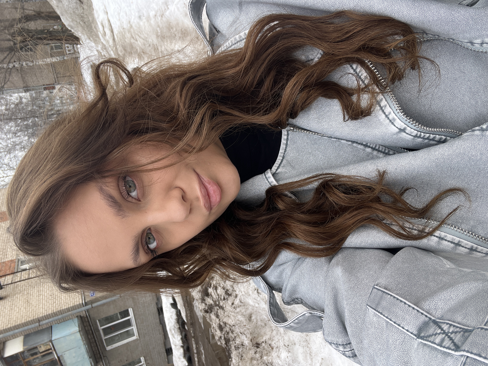 An outdoor, eye-level selfie captures a young adult female looking directly at the camera. She has long, wavy brown hair, green eyes, and light-toned skin, with a neutral expression. She is wearing a light grey, textured, leather-style jacket with a zippered front, over a black turtleneck garment.
The background on the viewer's left features a multi-story building constructed of light-colored brick, exhibiting white-framed windows and a light blue/teal enclosed balcony structure on an upper floor. A partial building number, possibly "12", is visible on the wall. To the viewer's right and behind the subject, a substantial pile of melting, dirty snow occupies the foreground, indicating winter or early spring conditions. Beyond the snow, leafless trees are visible, along with another distant building. The ground surface behind the subject appears to be a mix of melting snow and mud/slush. The scene is located in the city of Chaikovsky, Russia.