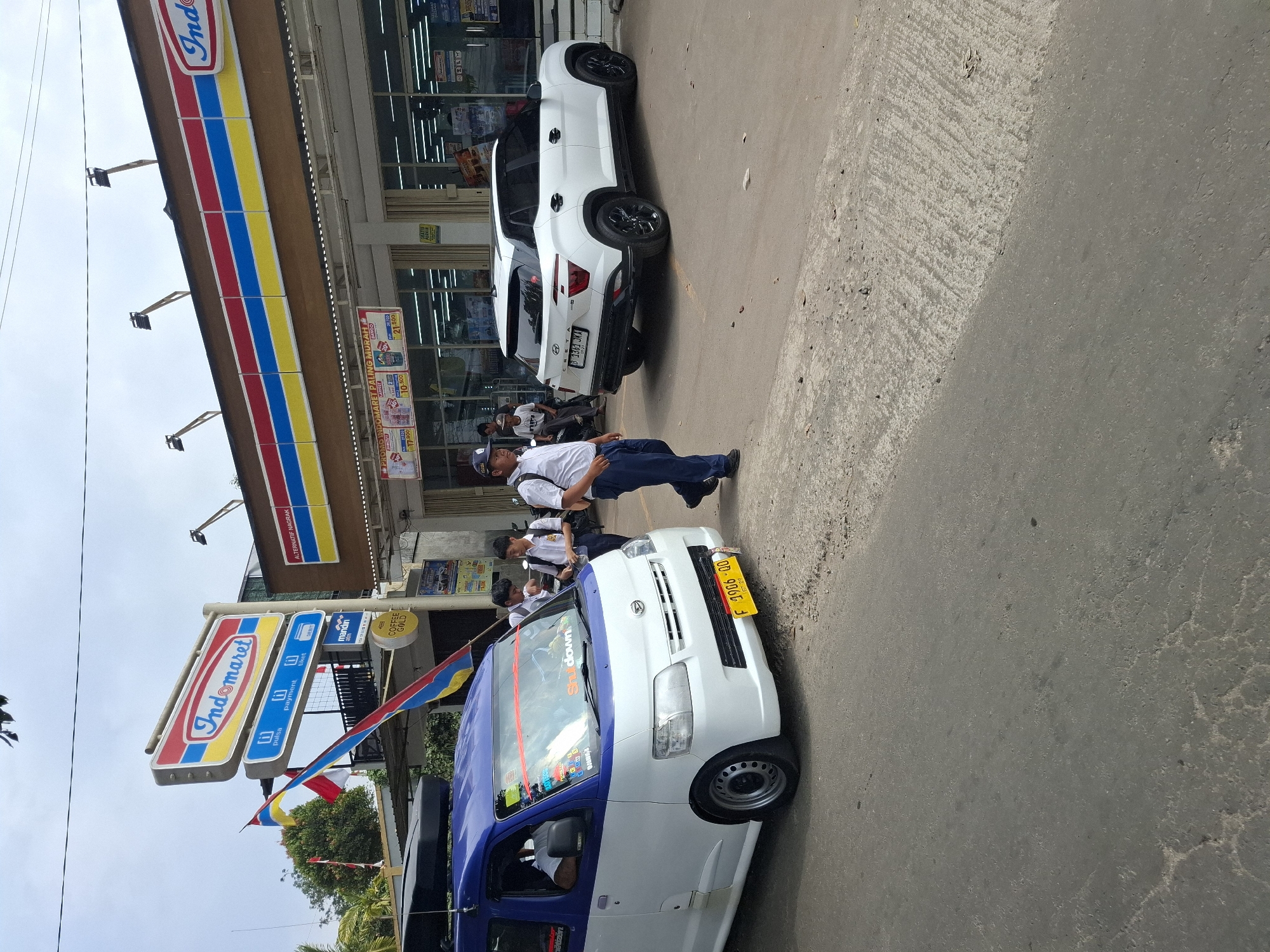 In Cibadak, Indonesia, on a cloudy day, a white and blue van with a roof rack is parked on the street in front of an Indomaret convenience store. The van has a yellow license plate that reads "F 1986 QD". Next to the van, a white Hyundai Creta SUV with black wheels is also parked. A man in a blue shirt and pants is walking away from the van towards the store. Several students in white shirts and blue pants with backpacks are walking on the sidewalk in front of the store. The Indomaret store has a prominent red, yellow, and blue sign, and promotional banners are visible in the windows. The street is paved with asphalt, and there is a concrete curb.