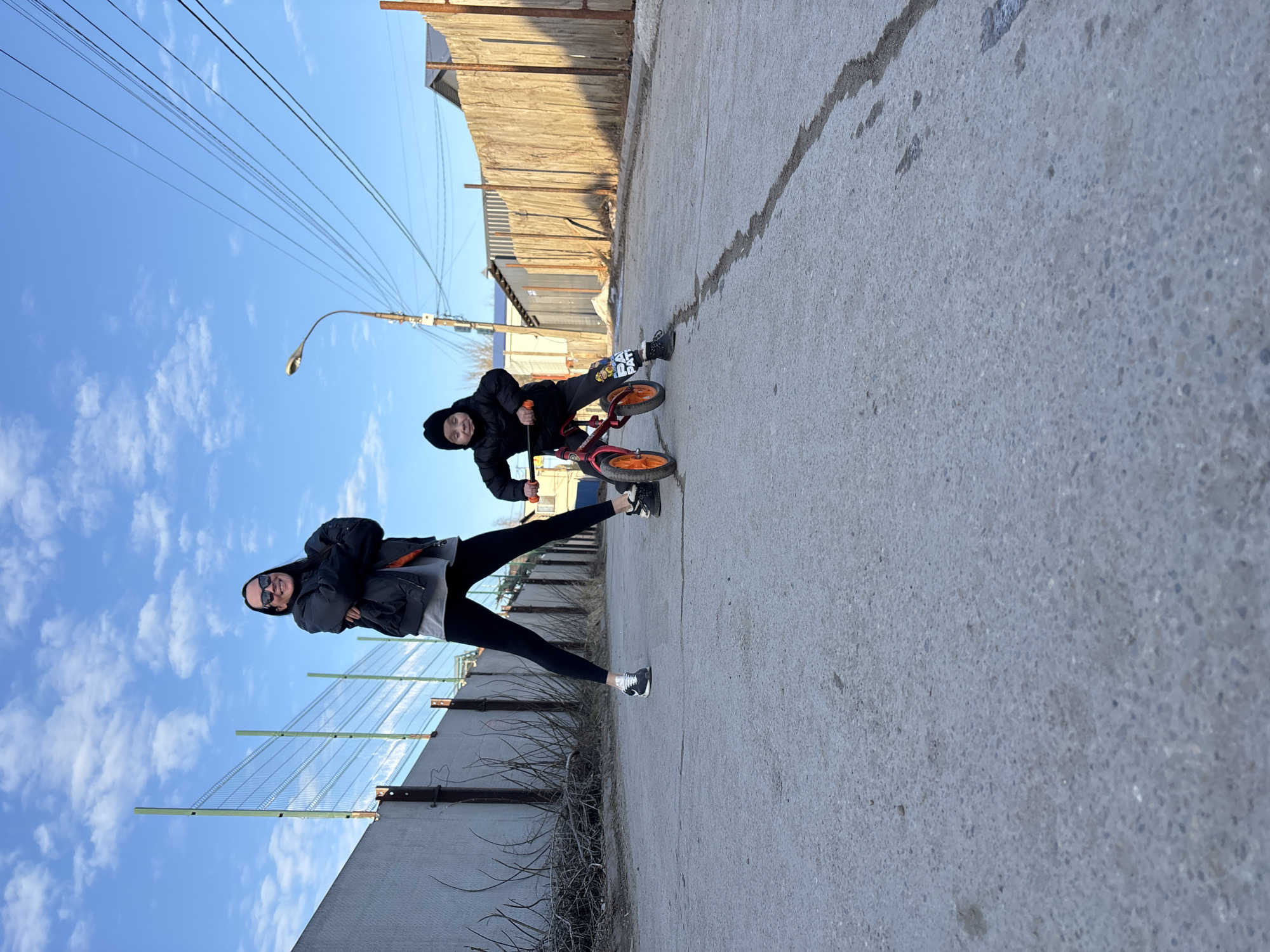 A photo shows a young woman and a child on a city street in Chaikovsky, Russia. The woman stands to the left wearing black leggings, black and white sneakers, a light gray top, and a dark jacket. She has sunglasses on and is smiling. To the right of her, a child is riding a small, red balance bike with orange wheels. The child is wearing dark clothing and a dark hooded jacket.
The street appears to be made of concrete. A metal fence runs along the left side of the street, and a wooden fence is on the right. A light pole is visible in the background, and power lines stretch across the blue sky with scattered clouds.
