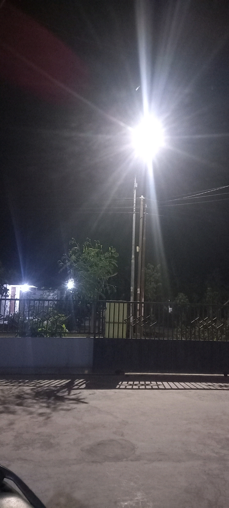 The image depicts an outdoor scene at night, illuminated by a bright streetlamp. The streetlamp is positioned to the right of the frame, casting strong light rays that cause lens flare. In the mid-ground, a dark metal fence runs horizontally across the image, with some greenery and structures visible behind it. To the left, a building with lights is partially visible. The foreground consists of a paved surface with shadows cast from the fence and possibly trees. The overall environment appears to be a residential or public area in the city of Pontang, Indonesia, with the streetlamp serving as the primary light source. There are no people or specific actions captured in the image.