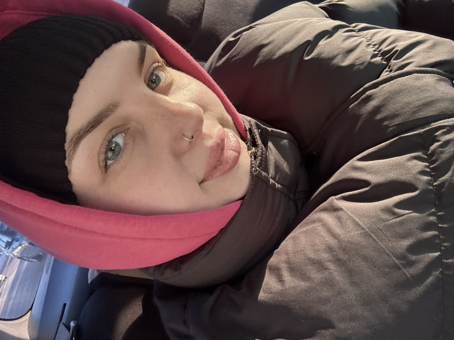 An image depicts a young adult female inside a vehicle, identified as being in Chaikovsky, Russia. The subject, viewed from the chest up, has light blue eyes, full lips with a subtle smile, and a small silver-colored hoop nose ring in her left nostril. She is wearing a black beanie, a bright pink hood over the beanie, and a dark, possibly brown or black, puffer jacket. Direct sunlight illuminates the left side of her face and jacket. In the blurred background, through a vehicle window on the upper left, other vehicles and a light-colored ground surface are visible. The interior of the vehicle behind the subject appears dark.
