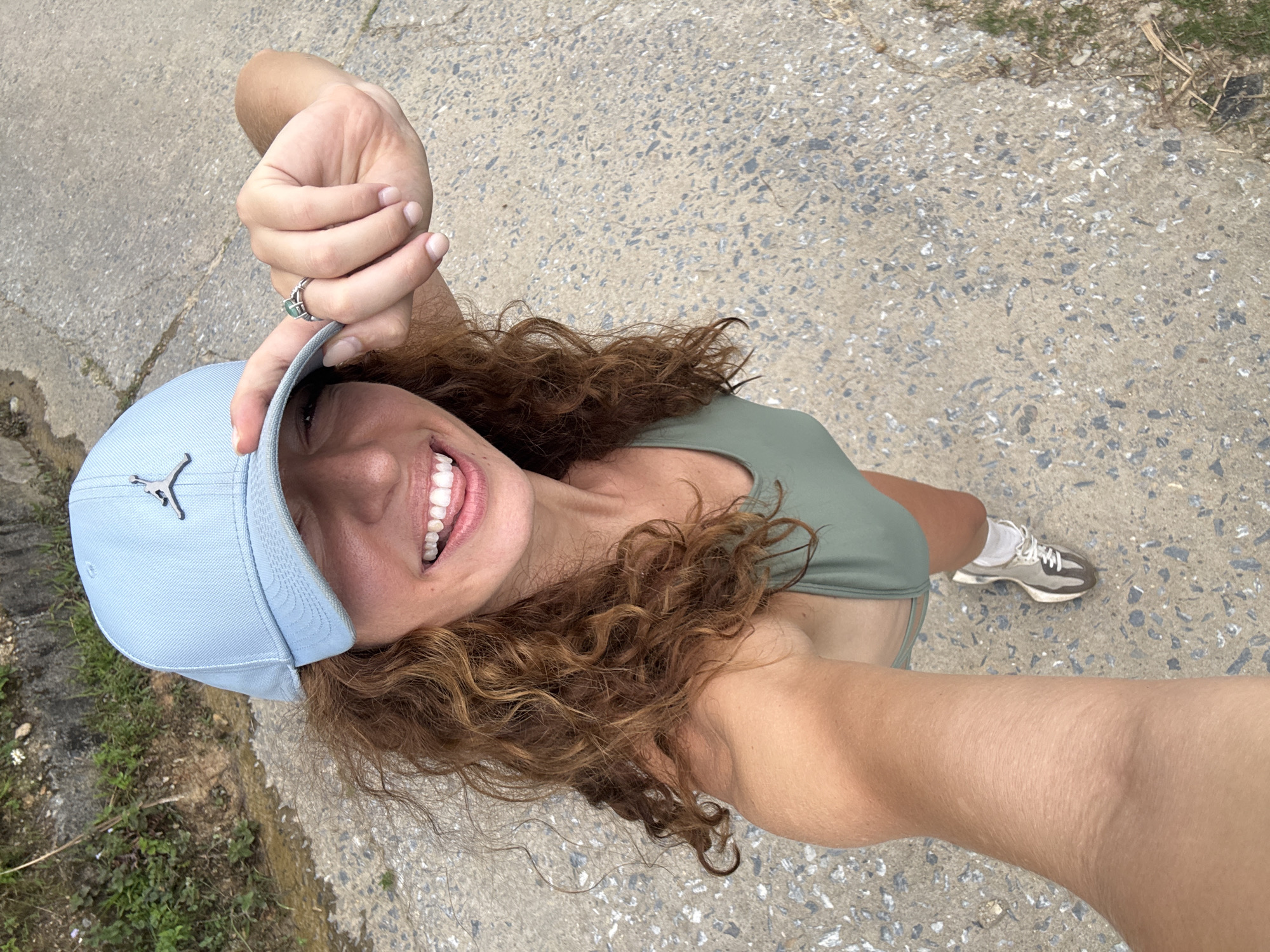 The image shows a woman in Suối Giàng, Vietnam, taking a selfie from a high-angle view. She is wearing a light blue baseball cap with a silver logo, a green top, and one visible running shoe. Her hair is curly and light brown. She is smiling, holding the brim of her cap with her left hand, which has a green ring on one finger. The background is a concrete path or road with some grassy areas visible along the edges.