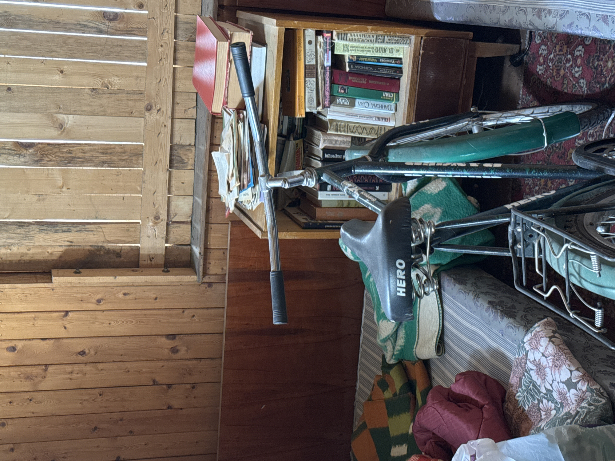 The image shows a cluttered indoor space. A wooden wall with horizontal slats is on the left. A bookcase filled with books is positioned near the wall. A bicycle is leaning against the bookcase, with its handlebars and seat prominently visible. The seat has the word "HERO" printed on it. A mattress lies near the bicycle. A floral patterned blanket is crumpled beneath the mattress. A wooden wall is behind the bicycle. A red item is laying near the blanket. The location and city are unknown.