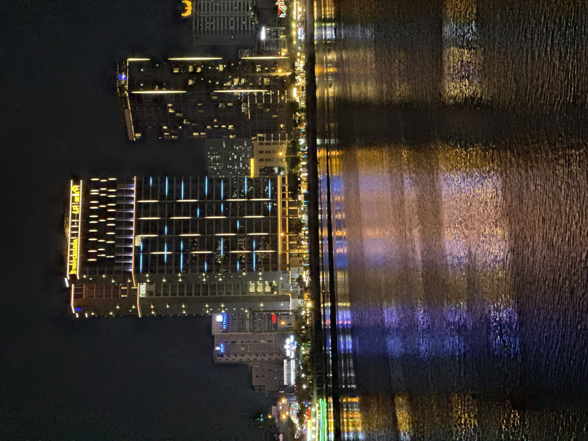 This media file presents a vibrant night scene along the waterfront of Đà Nẵng, Vietnam. The image is oriented vertically, with buildings on the left and the reflective surface of the water on the right.
The primary subjects are the illuminated city buildings and their reflections on the water. On the left, several multi-story buildings are brightly lit with various patterns of yellow, blue, and white lights, showcasing modern architectural designs. One prominent building features a yellow sign that appears to read "LA ANH" or "Lê Anh," accompanied by additional smaller text. Further along the waterfront, another building displays a recognizable "PETROLIMEX" sign, with what looks like a "KARAOKE" sign beneath it. In the upper part of the image (when rotated to landscape), a distinct yellow crescent moon-shaped light is visible on a building.
The activity is limited to the static display of urban life at night, with no explicit human figures visible. The setting is clearly a city at night, with artificial lights dominating the visual landscape.
Notable details include the striking reflections on the water, which are elongated and somewhat blurred due to the water's rippled surface, creating abstract streaks of yellow, blue, and purple light. A thin strip of land or promenade, also illuminated by numerous smaller lights, separates the buildings from the main body of water. The time of day is clearly nighttime, enhancing the dramatic effect of the city lights against the dark sky and water. The scene evokes a sense of urban energy and beauty after dark.