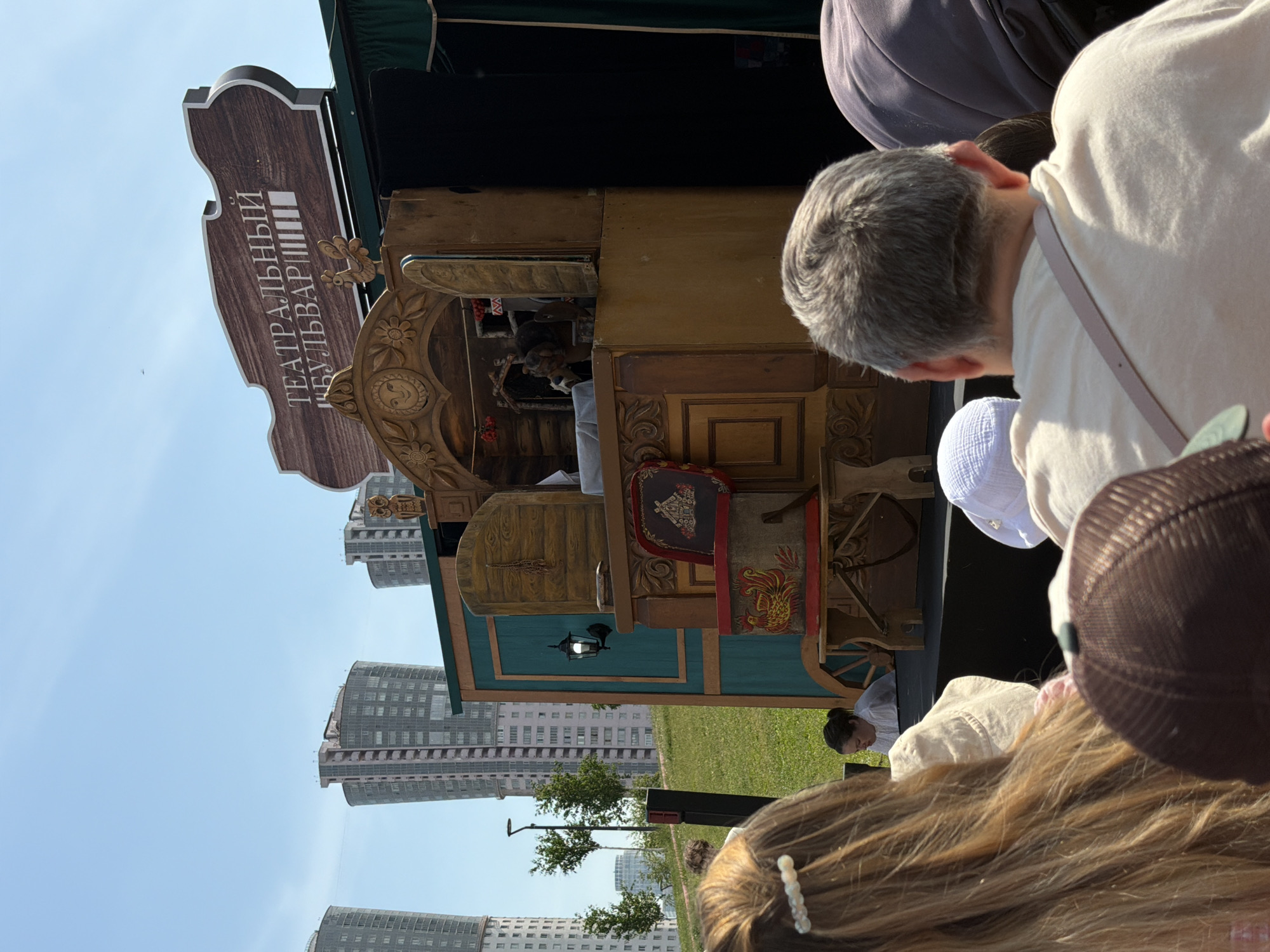 The image depicts an outdoor theatrical performance occurring on a sunny day in a park within the city of Khimki, Russia.
In the foreground, an audience is visible from behind, facing a wooden performance booth. To the immediate left, the back of a person with long, light brown hair, adorned with white pearl clips, is seen. To the immediate right, the back of an adult with short, graying hair, wearing a light-colored t-shirt with a light pink strap over the shoulder, is present. Adjacent to this individual, a small child's head with a white hat and part of a baby stroller are visible.
The central subject is a freestanding, ornate wooden performance booth, likely a puppet stage, featuring intricate carvings, including a sun face at the top center of an archway and a small rooster figure atop the right side. The booth incorporates teal-colored panels with wooden trim and a decorative wall lamp on its left exterior. Inside the arched opening, puppets or figures are partially visible, one appearing animal-like, alongside decorative elements resembling red berries. A red-painted chair or box with a stylized rooster design is positioned in front of the lower section of the booth.
Above the performance booth, a prominent, dark brown wooden sign with white lettering displays "ТЕАТРАЛЬНЫЙ БУЛЬВАР" (Theatrical Boulevard) in Cyrillic.
The mid-ground features a gently sloping green grassy area. A single adult with dark hair tied back, wearing a white shirt, is seated on this incline, observing the performance. Several small deciduous trees and a black lamppost are also situated on the grassy slope.
The background is dominated by a clear, light blue sky and multiple modern, tall residential high-rise buildings, characterized by numerous windows and varying architectural shapes, suggesting an urban environment.