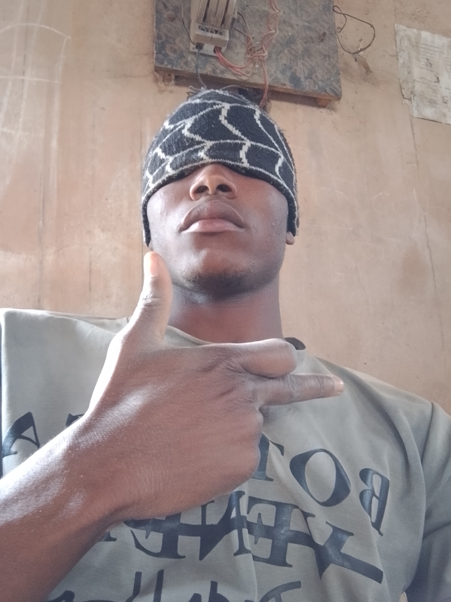 This is a close-up, waist-up selfie of a young Black man wearing a black and white patterned beanie that covers his eyes, obscuring his face. He is wearing a light gray t-shirt with large, dark, bold lettering. His right hand is raised, and he is making a gesture with his thumb extended upwards in a "thumbs up" sign, and his index and middle fingers extended outwards in a "peace sign" gesture. His skin tone is dark. The background appears to be a wall made of plastered earth or concrete, with some visible electrical wiring and a small electrical box mounted high on the wall. There are some faint chalk markings on the wall to the left of the subject. The lighting suggests it could be daytime. The overall impression is casual and posed.
The text on the t-shirt appears to be in bold, dark letters and is partially obscured by the man's hand and the angle of the shot. The visible letters include "BO" and "Y", suggesting words like "BODY" or similar. The setting seems to be indoors or a sheltered outdoor area, possibly a residence in Bukuru, Nigeria, given the style of the wall and electrical fittings. There are no explicit indications of weather conditions or a specific time of day beyond the general lighting. The subject's pose and gesture convey a sense of positivity or affirmation.