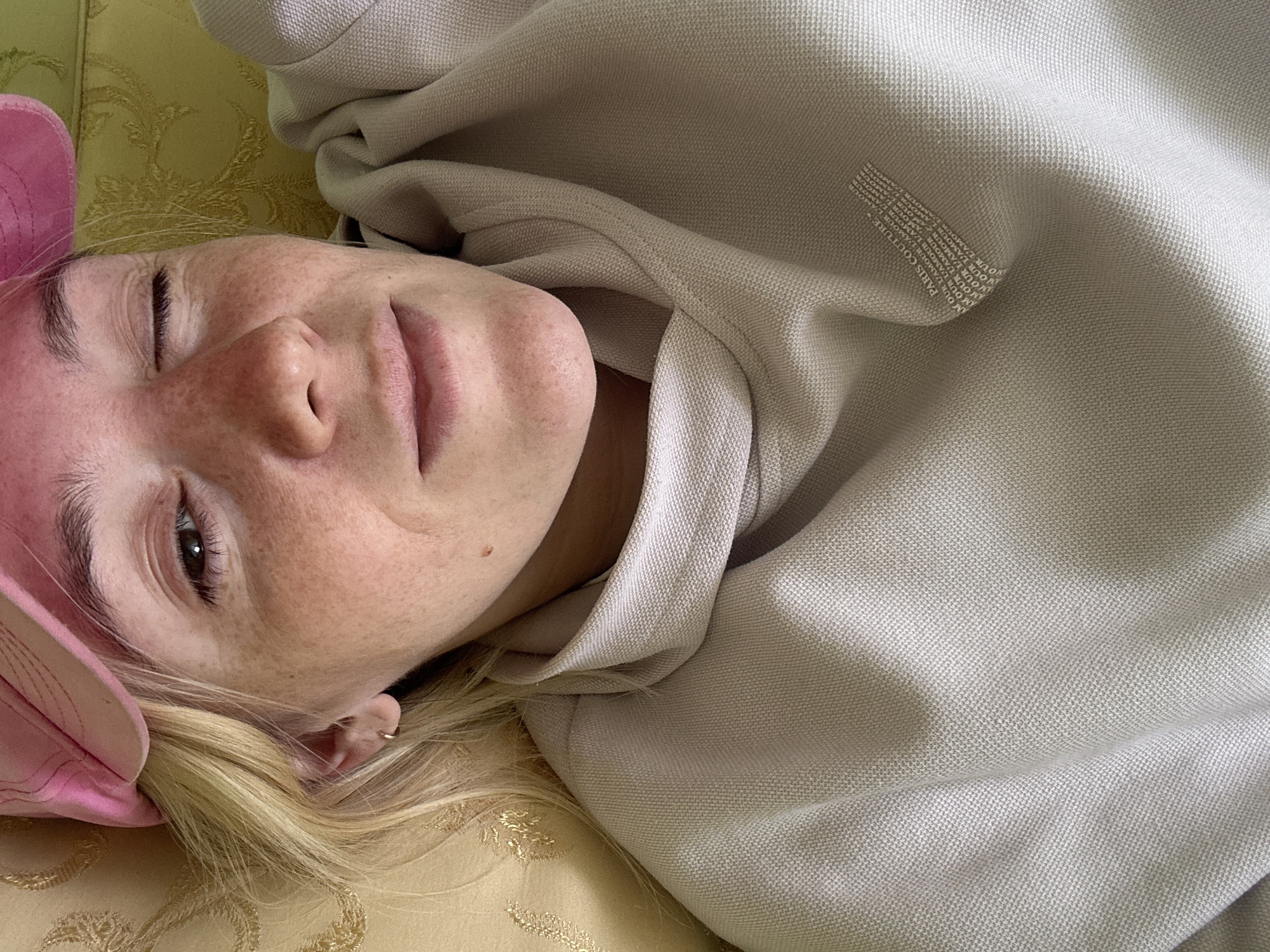 A close-up, overhead shot shows a person with blonde hair and freckles wearing a pink hat and a beige hooded sweatshirt. The person is lying down, propped up by a yellow patterned cushion. They are looking at the camera with one eye open and the other closed, with a slight smile. The sweatshirt has a repeating text logo on the chest that reads "PARIS COUTURE AMOUR L'AMOUR". The background is a yellow patterned fabric, possibly a couch or armchair. The image appears to have been taken indoors, in a well-lit room. There is no indication of the location being in Sarov, Russia.