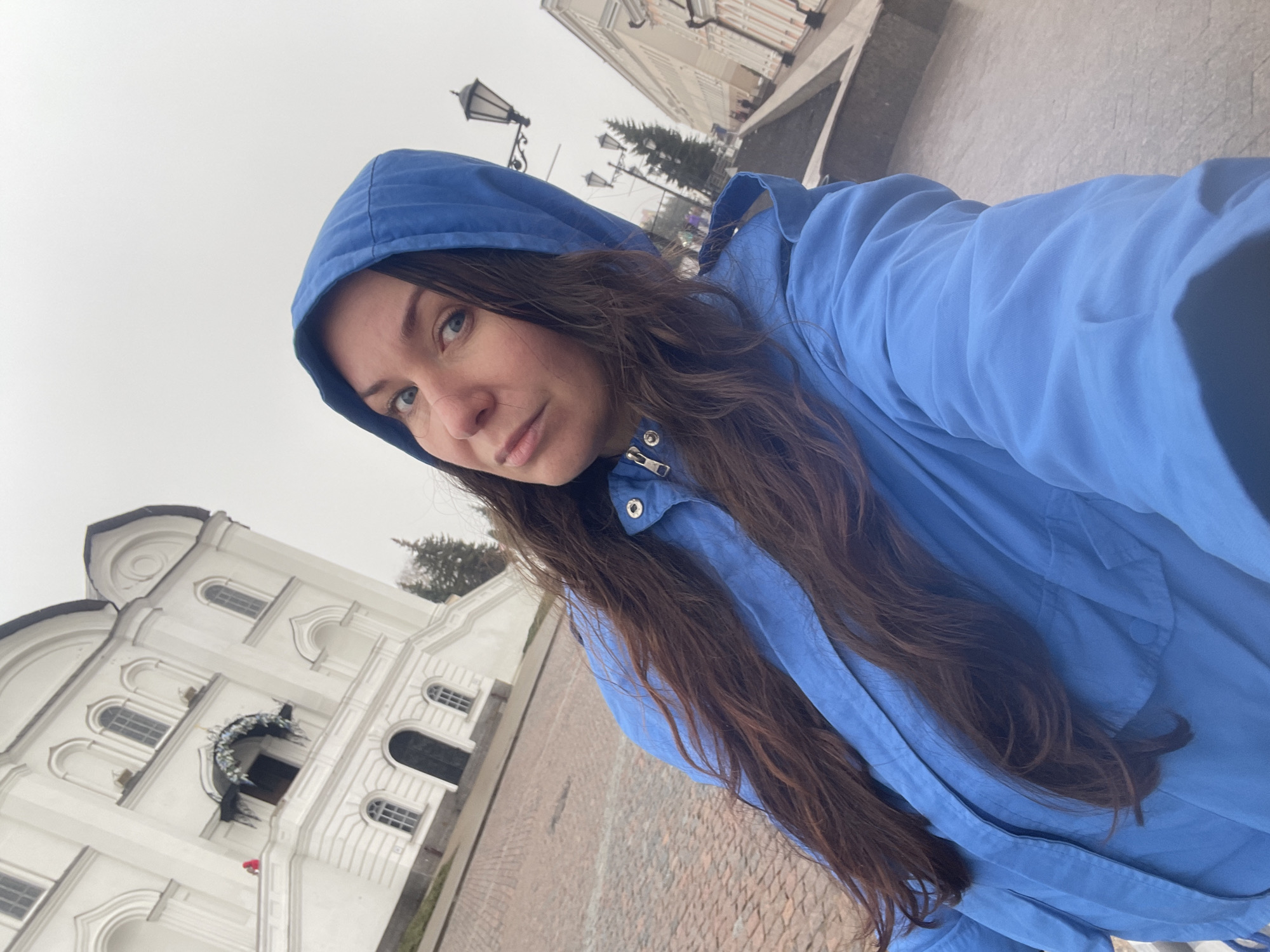 A woman wearing a blue hooded jacket poses for a selfie in Kazan, Russia. In the background, a white multi-story building with arched windows and decorative wreaths above its entrances is visible to the left of the woman. Stone paving covers the ground. A streetlight stands tall, with several other buildings and trees behind the woman. The sky is overcast.
