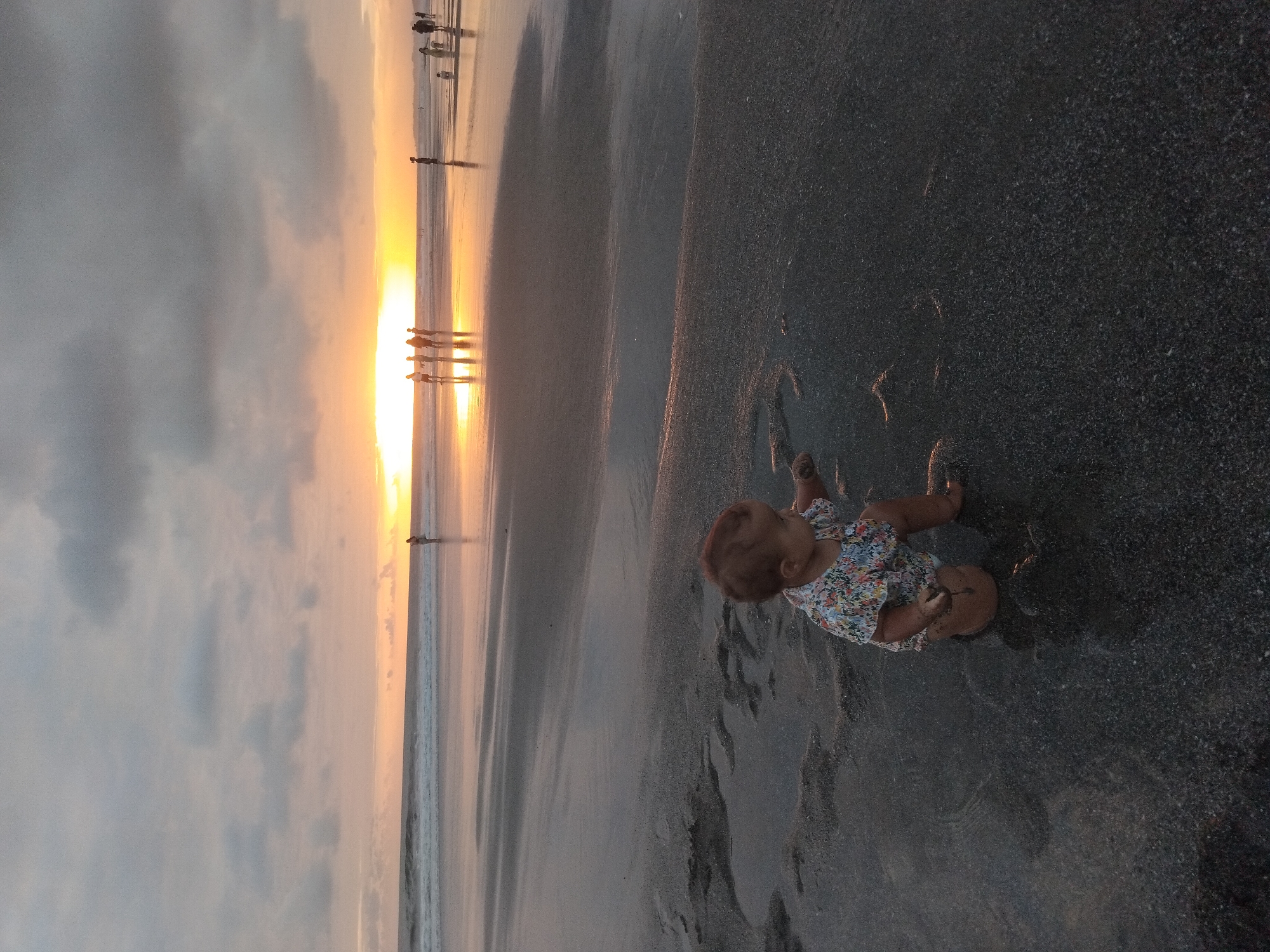 A baby wearing a floral print shirt and diaper kneels on a wet, dark sand beach. The baby is playing with sand, holding some in its right hand. In the background, the ocean meets the horizon under a cloudy sky with a bright sunset. Several figures, appearing to be adults and children, are silhouetted against the sunset in the distance. The location is identified as the beach in North Kuta, Indonesia.