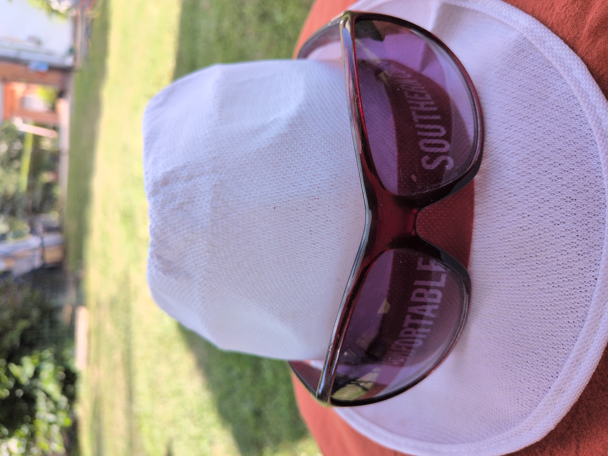 A white bucket hat with sunglasses is placed on an orange surface. The sunglasses have purple lenses and a red frame. Text is visible on the lenses, reading "COMFORTABLE" on the left lens and "SOUTHERN" on the right lens. The background is blurred and shows a green lawn with trees and a building. The location is the city of Rastenberg, Germany.