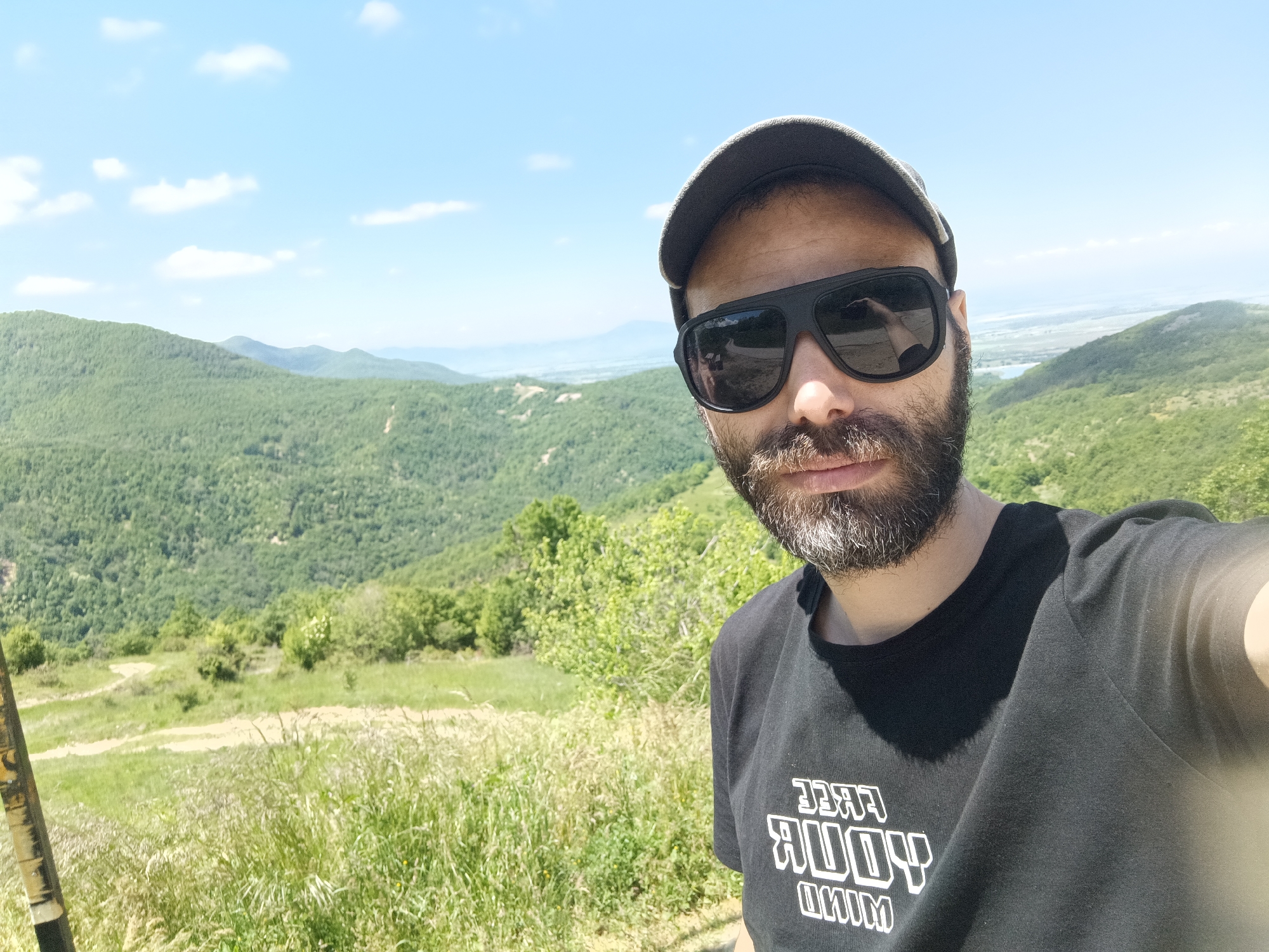 A man with a beard and sunglasses, wearing a cap and a black t-shirt with white writing, is taking a selfie. The background shows a hilly landscape with green vegetation under a blue sky with scattered clouds. The location is Oreshets, Bulgaria.