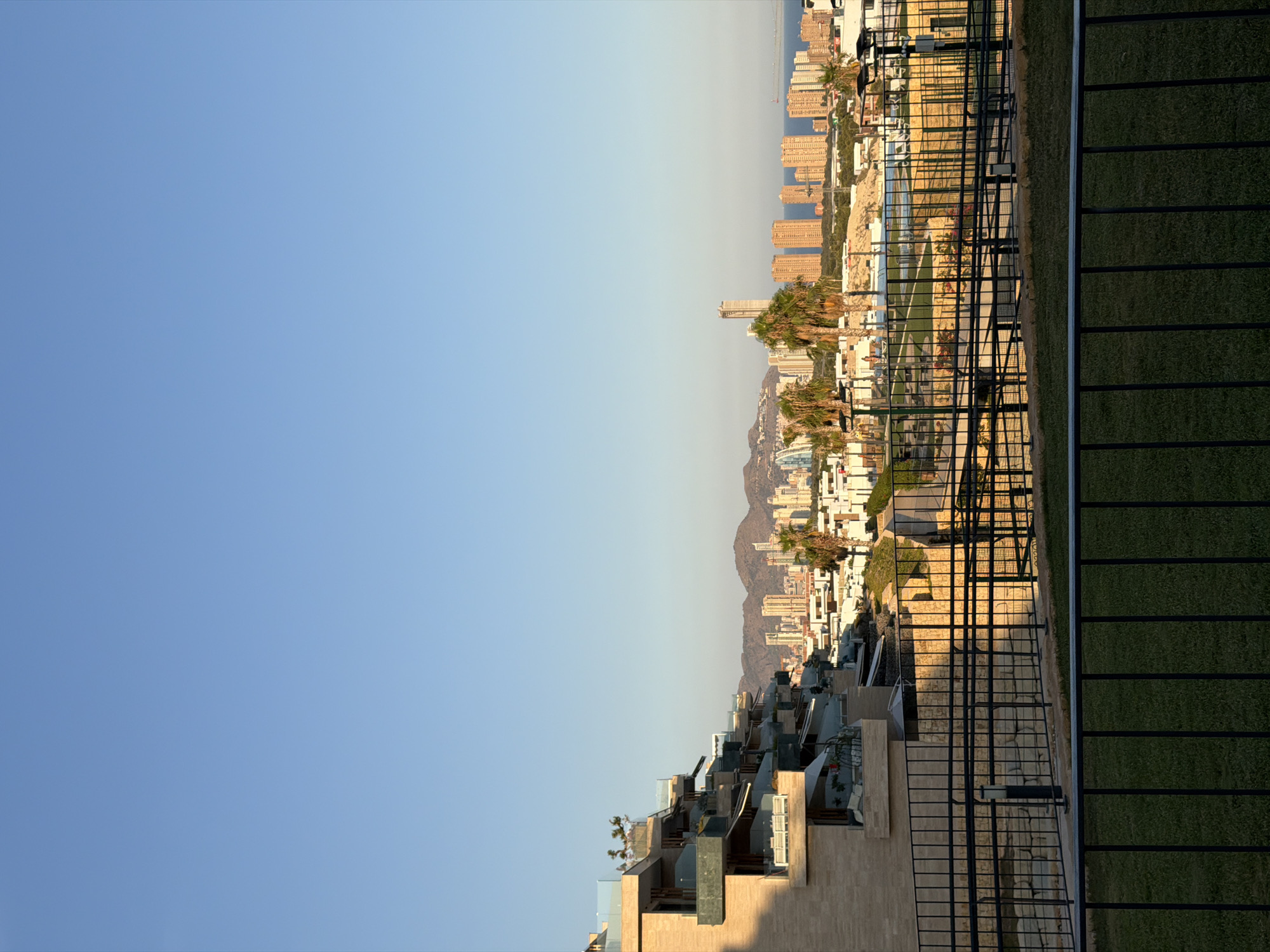 This image shows a view of the skyline of Finestrat, Spain, from a residential building. The foreground features a modern building with stepped balconies, complete with outdoor furniture. A black metal fence runs across the mid-ground, behind which is a grassy area with more fencing. Beyond this, a cityscape unfolds with numerous tall buildings and palm trees. In the distance, mountains form a backdrop, and the sea is visible under a clear blue sky. The sun is low in the sky, casting long shadows and illuminating the buildings with a warm light. There are no people visible in the image.