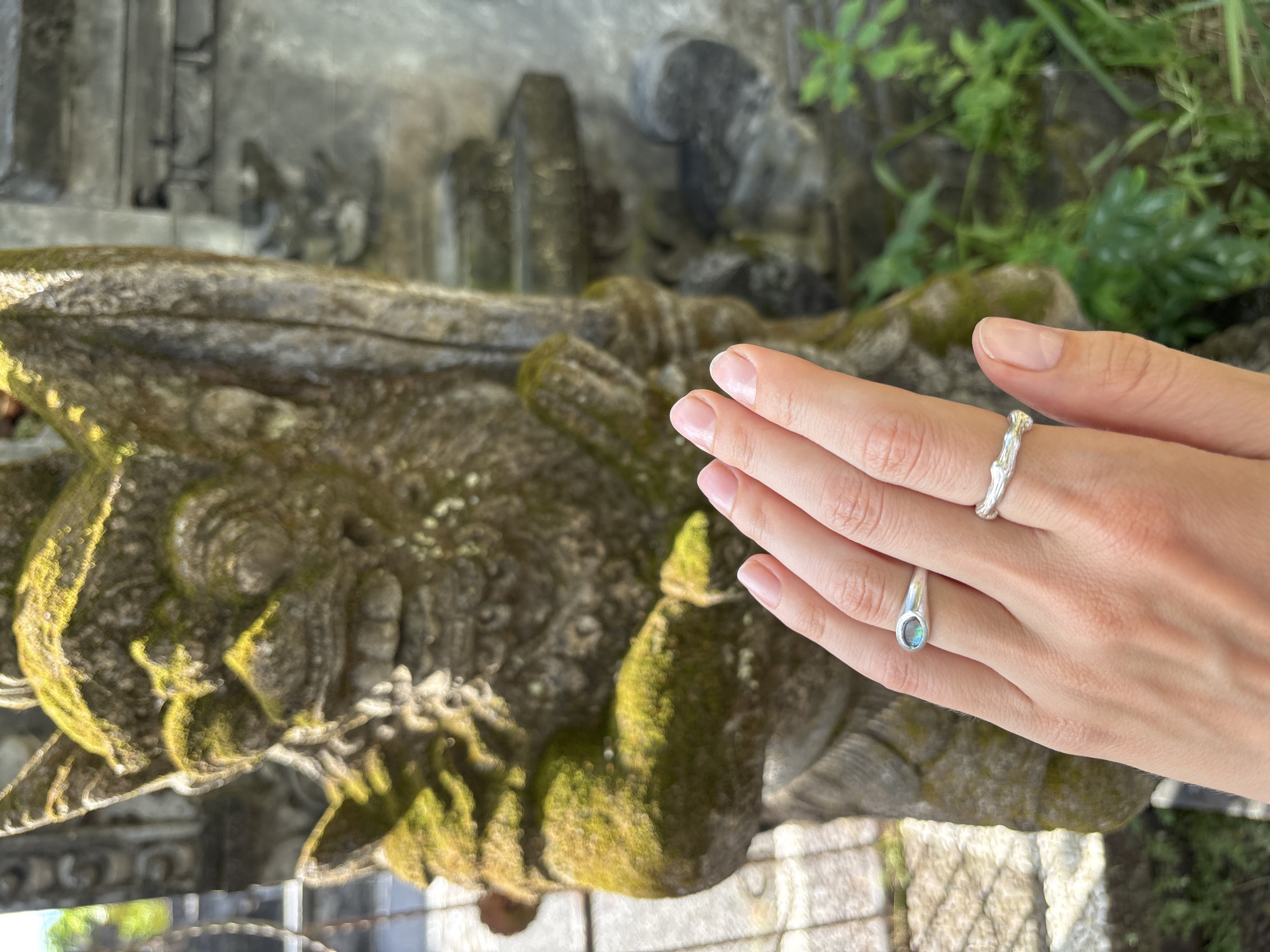 The image captures a close-up view of a person's hand adorned with two silver rings. One ring, worn on the index finger, has a nature-inspired design resembling a twig or branch. The other, on the middle finger, features an oval stone with iridescent hues. The hand is positioned against a moss-covered stone sculpture or architectural detail. The background includes foliage. The location is Kuta Selatan, Indonesia.