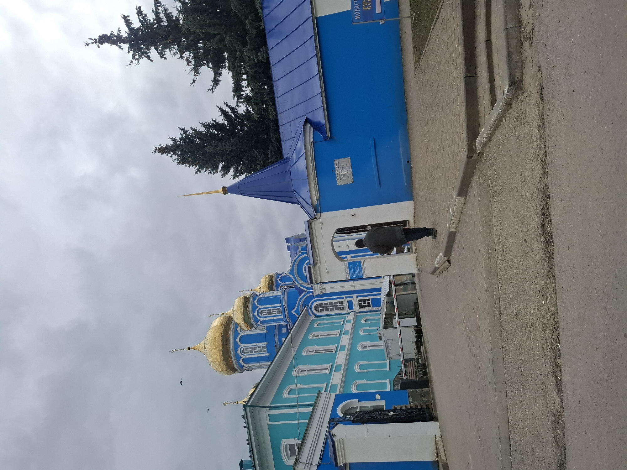 This image captures a street-level view of a vibrant Russian Orthodox monastery complex in Zadonsk, Russia, under an overcast sky.
The scene is dominated by a collection of religious buildings, characterized by their striking blue and white facades and multiple golden onion domes, typical of Russian Orthodox architecture. The primary church building is adorned with several large, gleaming golden domes and crosses, intricately detailed in blue and white patterns. Adjacent to it, a lighter blue building with white trim adds to the complex. Further back and partially obscured by trees, another deep blue building with a matching blue sloped roof and a small golden spire is visible.
A single person, seen from behind, dressed in a dark jacket, walks along a patterned brick sidewalk towards the monastery buildings. To the right of the sidewalk, an asphalt road runs parallel. The ground also features concrete curbs and drainage elements.
The sky is uniformly overcast, filled with light grey clouds, suggesting a cloudy day, possibly late morning or afternoon given the ambient light. Dense, dark green coniferous trees frame the upper parts of the image, contributing to the serene atmosphere.
A notable detail is a white sign with blue text affixed to the deep blue building. The visible Cyrillic script clearly reads "МОНАСТЫРЬ" (Monastery). Below it, partially obscured, are the letters "БУХ...", likely part of "Бухгалтерия" (Accounting) or a similar designation. The overall impression is one of a peaceful, everyday scene at a significant religious site.