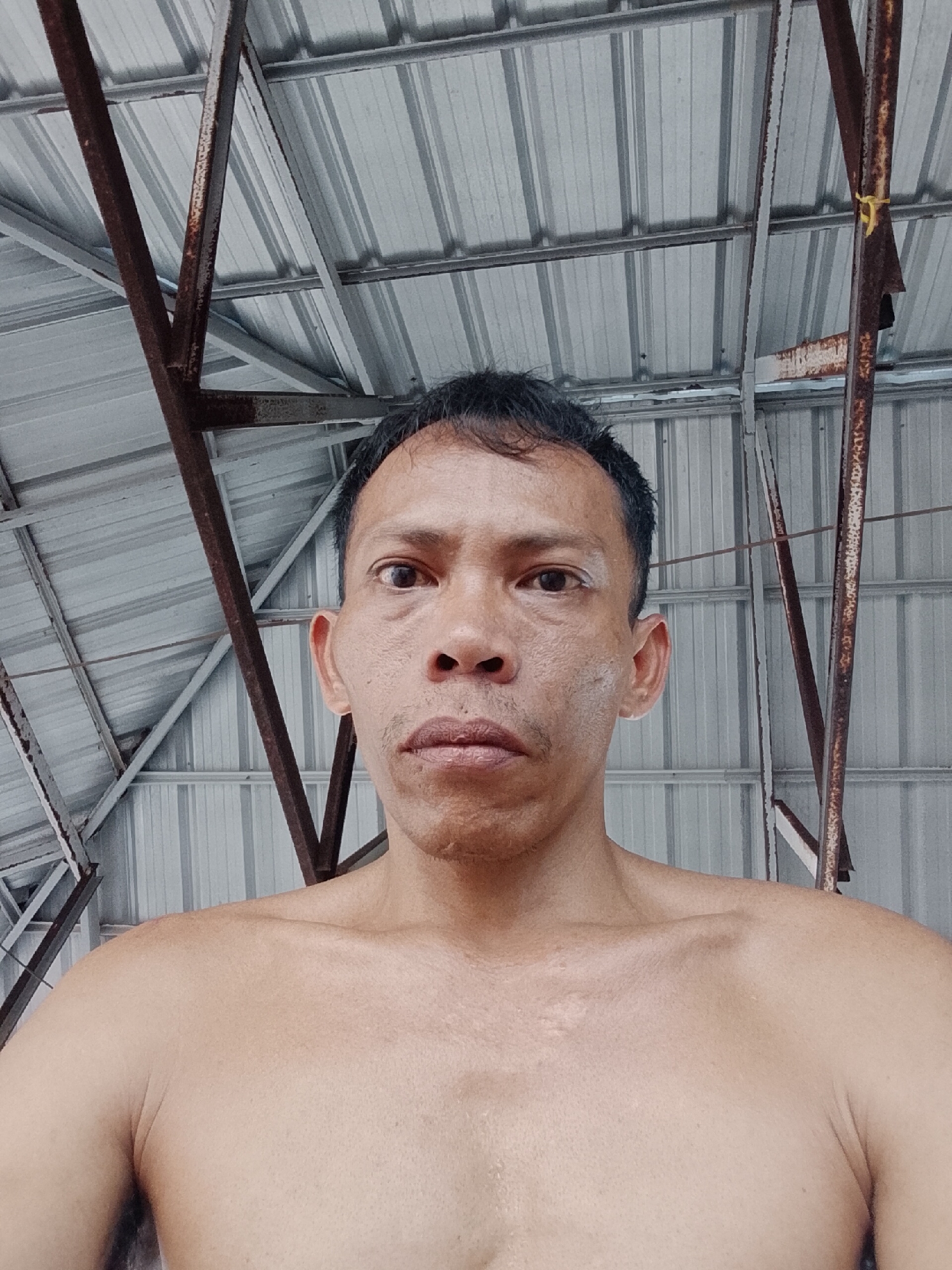 A medium shot depicts a middle-aged Asian man with dark hair and a serious expression. He is shirtless and has skin imperfections on his face. The background shows a corrugated metal roof with rusty metal supports, suggesting an outdoor or semi-enclosed structure. The location is Bukit Ketok, Indonesia.