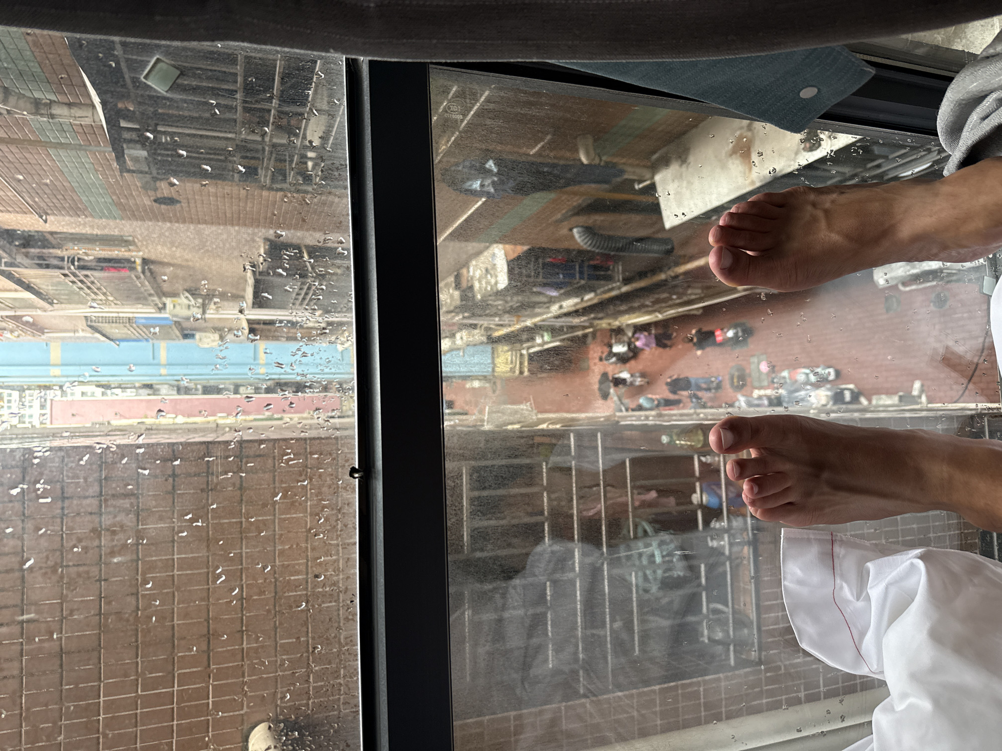 The image is a perspective shot looking down through a rain-streaked window from a high vantage point, seemingly inside a building. Two bare feet are visible in the foreground, resting near the window's edge, indicating someone is looking out. A white cloth is draped nearby, suggesting the presence of bedding or similar fabric.
Outside the window, a cityscape is visible, with buildings of varying heights and colors. The overall scene is somewhat obscured by the rain and the angle of the shot, but the arrangement of buildings and urban elements suggests a densely populated area. Some people appear to be in the public spaces below.
Based on the details visible in the architecture and cityscape, it is suggested to be Futian District, China. However, it cannot be confirmed with certainty without further context.
