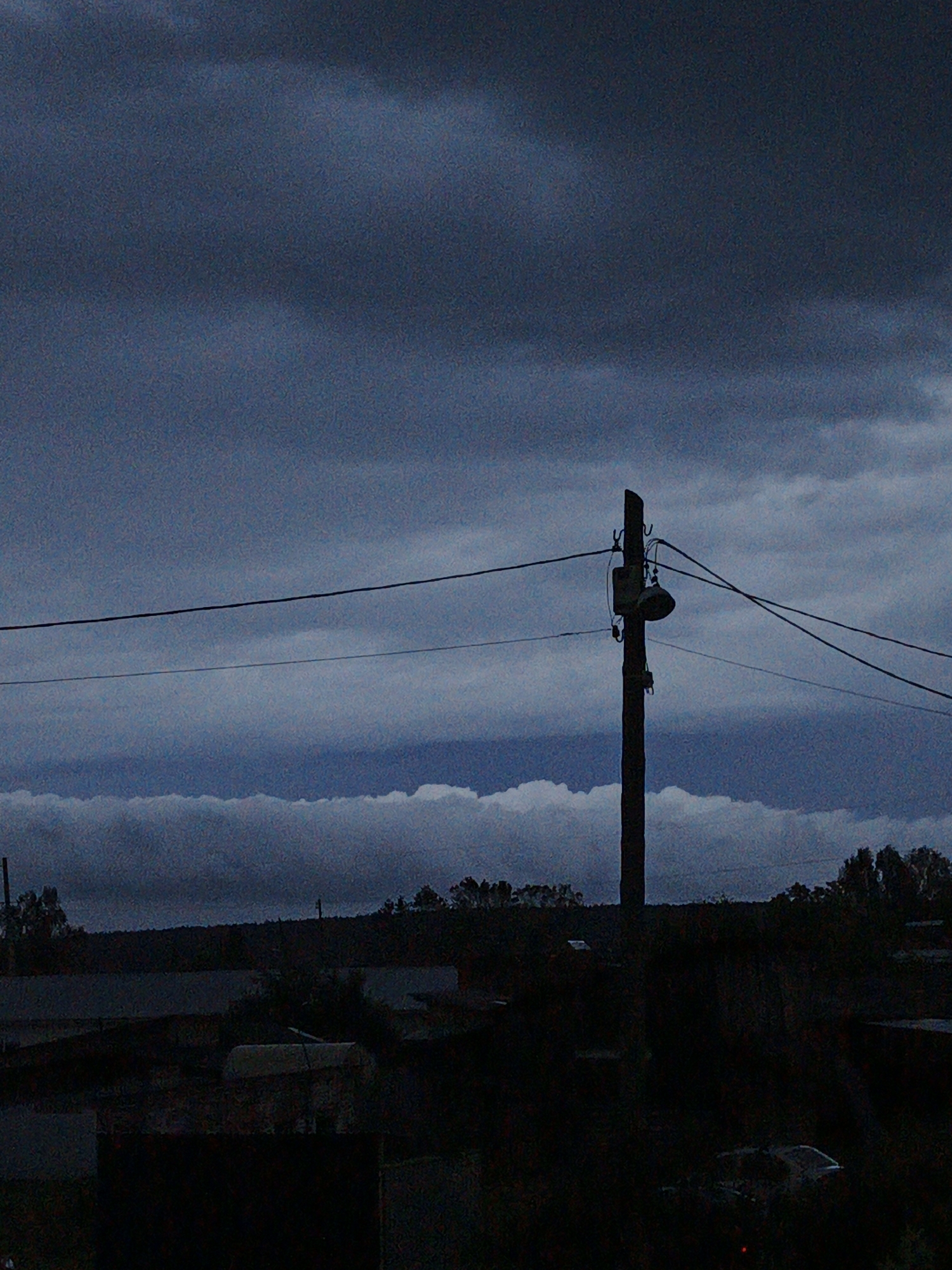 A dark, grainy photograph captures a utility pole with a light fixture silhouetted against a cloudy, twilight sky. The pole stands in the foreground, with wires extending horizontally across the frame. Below the pole, a dimly lit landscape shows the dark shapes of buildings and trees, suggesting a rural or semi-urban setting. The overall mood is somber and quiet, possibly indicative of approaching night or inclement weather.