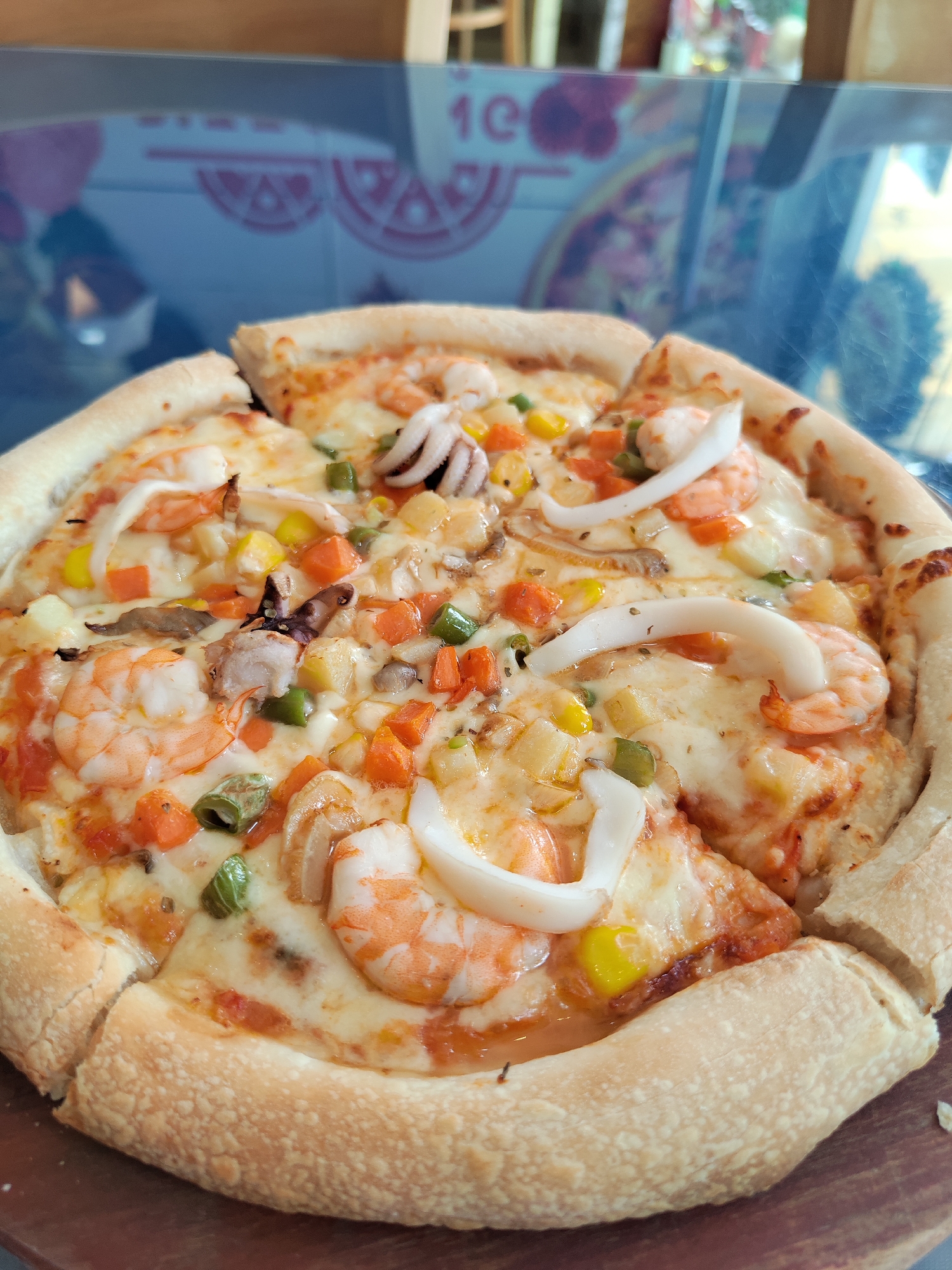The image shows a seafood pizza on a wooden board. The pizza is topped with shrimp, squid rings, corn, diced carrots, pineapple, green beans, and cheese. The crust is thick and golden brown. The pizza is cut into slices. The background is a blurry reflection of what appears to be a menu board in a restaurant. Based on information available online, the picture was taken in Da Lat, Vietnam.
