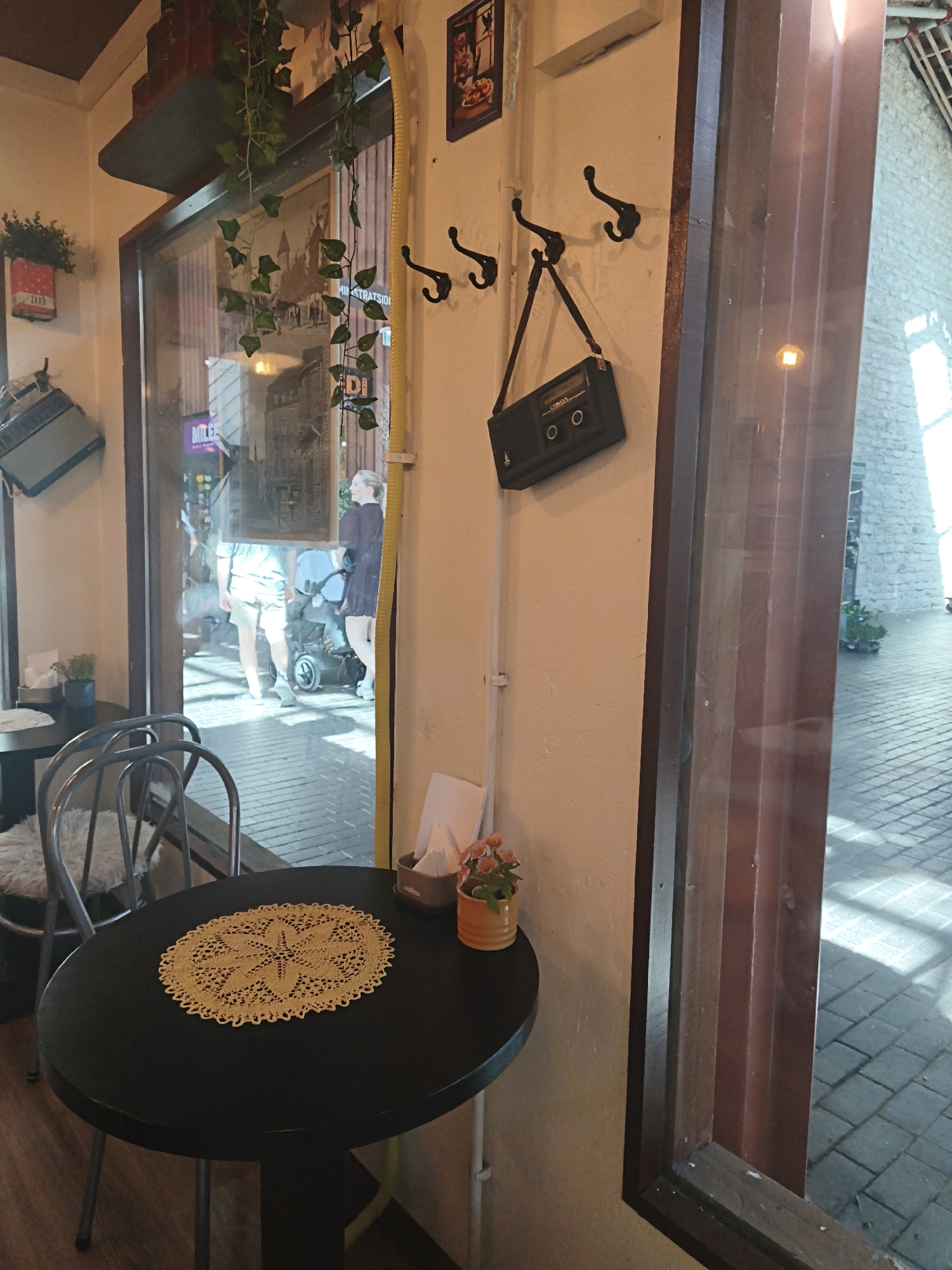 This image depicts the interior of a cafe, likely located in Tallinn, Estonia, given the reflection in the window which shows people walking outside in what appears to be a European-style urban setting. In the foreground, a small, round, black table is set with a decorative yellow doily and a small potted plant with orange flowers. To the left of the table, a metal chair with a white cushion is visible. The wall behind the table is cream-colored and features several hooks with a black retro-style radio hanging from one. A yellow electrical conduit runs vertically along the wall. Through a large window, which forms the right side of the image, a sunlit outdoor area with patterned pavement and a stone building is visible. The reflection in the window shows pedestrians, one of whom is pushing a stroller. Green ivy plants hang from a shelf above the window.