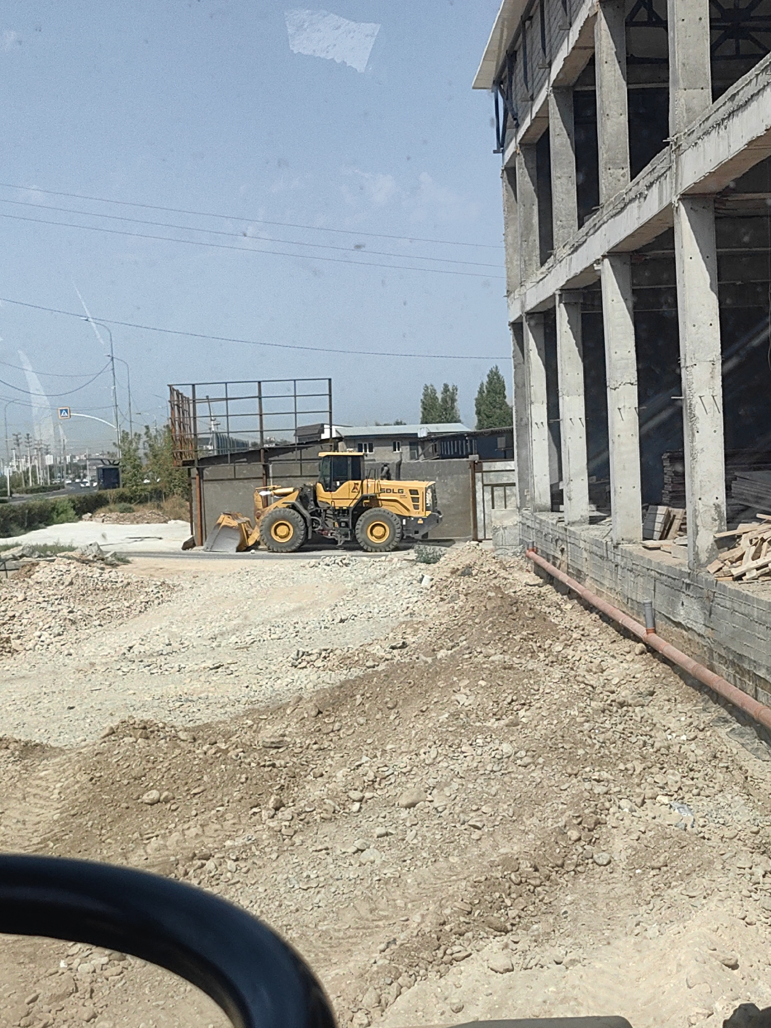 The image depicts a construction site in the city of Shymkent, Kazakhstan. In the foreground, a yellow front-end loader manufactured by SDLG is parked on a gravel and dirt surface. The loader has its bucket lowered to the ground. Behind the loader, a partially constructed concrete building dominates the right side of the frame. The building features large concrete pillars and beams, with open-air floors and exposed rebar. To the left of the loader, a metal fence structure is visible. In the background, a street with traffic and power lines suggests an urban environment. The sky is clear and blue. The overall scene suggests ongoing construction work in an industrial or developing area of Shymkent.