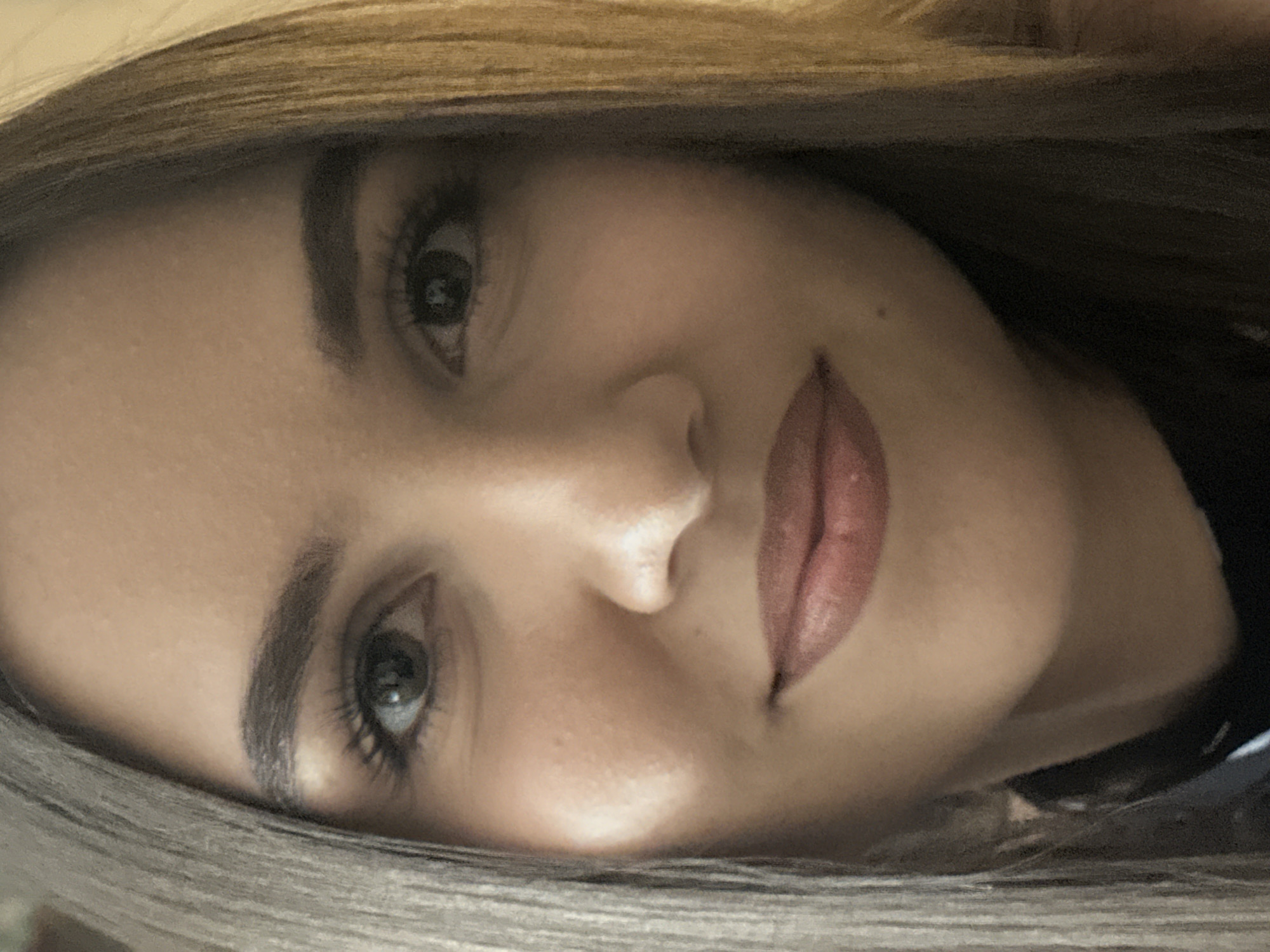 A close-up, slightly angled portrait captures the face of a young woman. Her light brown eyes are open and looking forward, framed by dark eyebrows and long eyelashes. Her lips are slightly parted, and she appears to be wearing lipstick. Her hair is long and appears to be a mix of brown and blonde tones, falling around her face. The background is indistinct and dark. The image was taken in an indoor setting.
