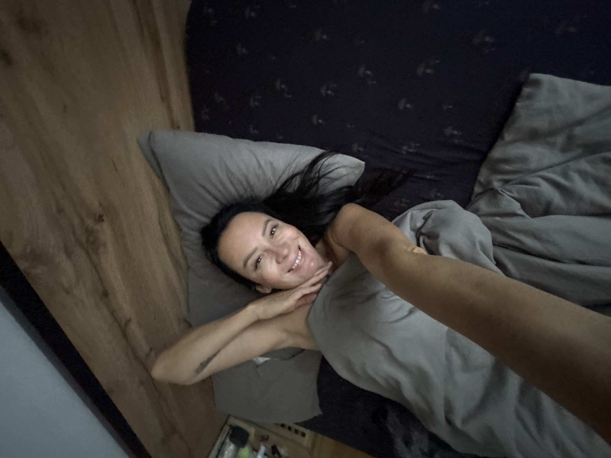 A woman with dark hair and brown eyes is lying in bed, looking at the camera and smiling. She is wearing a gray tank top and is covered by a gray blanket. Her left arm is extended towards the camera, and her right hand is resting on her neck. The bed has a dark patterned sheet and a gray pillow. To the left of the bed is a wooden floor. The scene appears to be a bedroom, possibly in Chaikovsky, Russia.