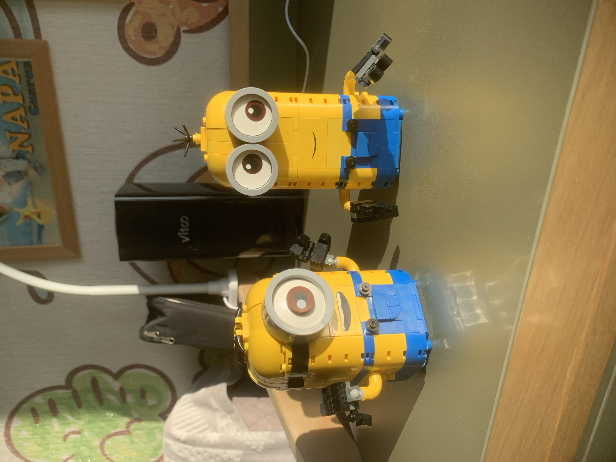 A high-angle shot depicts two Lego Minion figures on a surface that appears to be a desk or tabletop. The figures are predominantly yellow with blue accents, and have characteristic single or double eyes. One Minion has its arm raised, equipped with a mechanical-looking claw or tool. The figures are positioned near a black rectangular object with the brand "Vita" visible on it, possibly a power bank or similar electronic device. A white cable connects to the device, extending towards the lower left of the frame. The surface beneath the Minions is a light-colored material with a patterned design that includes circular and cloud-like shapes. A partially visible picture frame is present in the upper left corner. The right side of the frame suggests the presence of a wall or vertical surface. Based solely on the visual evidence, there's no data to confirm the location of the image as being Izhevsk, Russia or any specific city. The scene is brightly lit, suggesting daylight conditions.