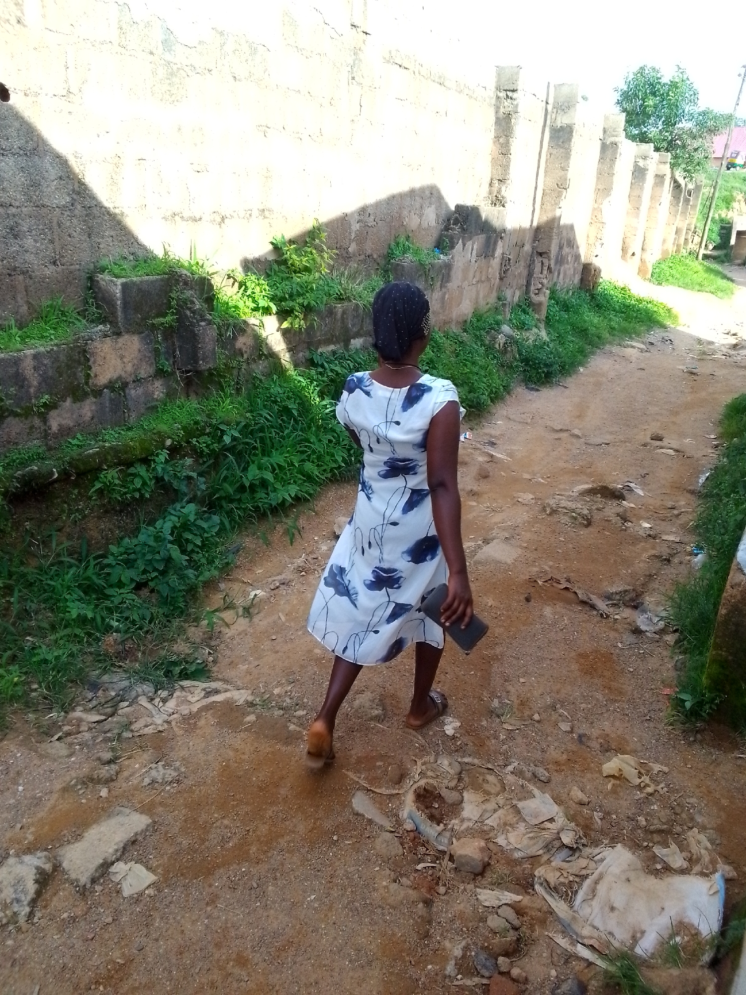 A rear view of a woman with dark skin and dark hair, wearing a white dress with a blue floral pattern, walks down a dirt path. She is holding a dark-colored rectangular object in her right hand. The path is lined on the left by a rough, stone wall with overgrown green vegetation. On the right, the path continues into the distance, with more green vegetation and a hint of a building in the background. The ground is uneven and appears to be a mix of dirt, gravel, and some debris. The lighting suggests it is daytime, with strong shadows cast by the wall.