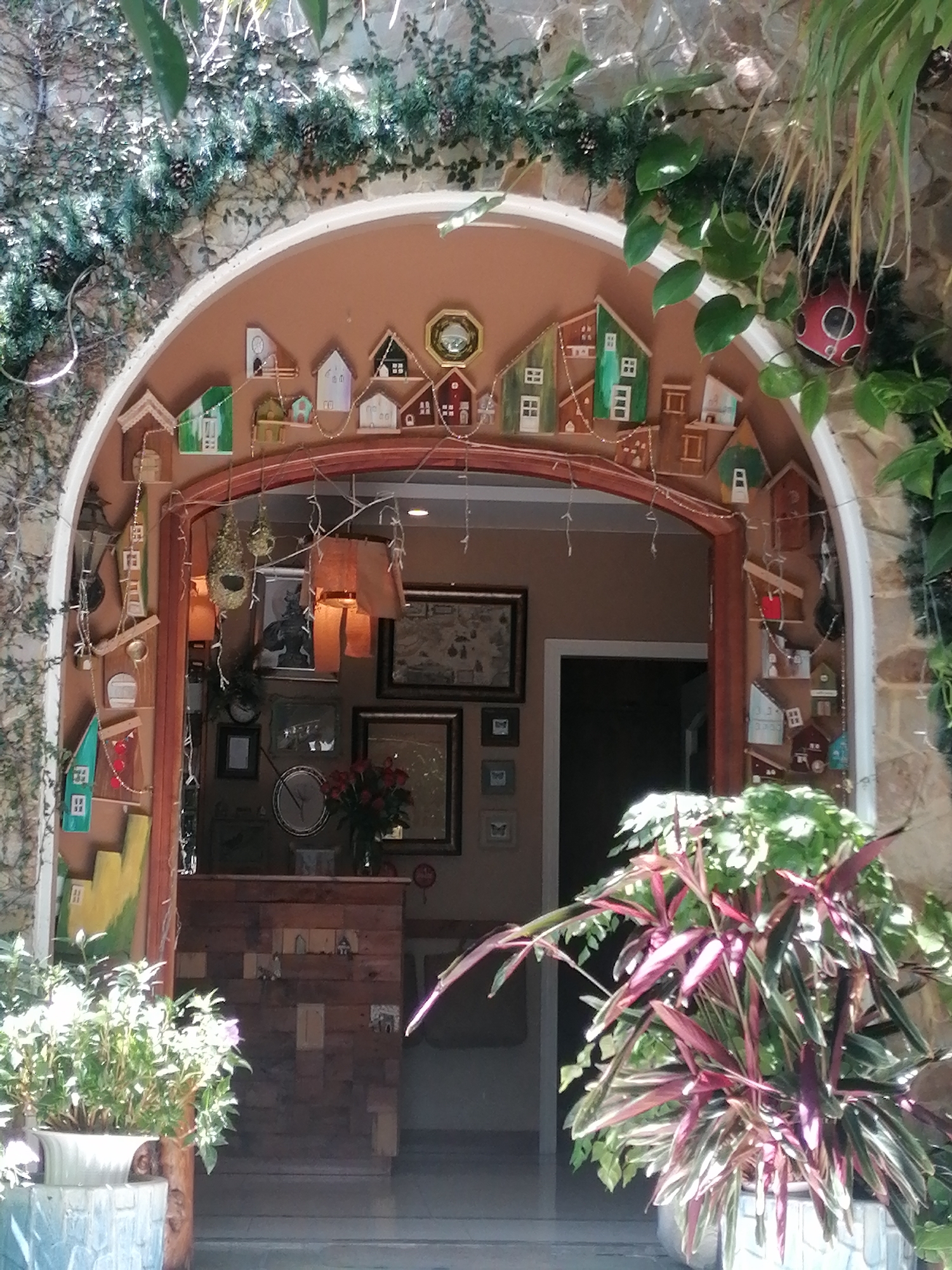 The image shows the arched entrance to what appears to be a quaint cafe or shop in Da Lat, Vietnam. The entrance is adorned with an abundance of small, decorative wooden houses, strung together with fairy lights and tinsel, suggesting a festive or whimsical theme. A stone wall and lush green foliage frame the upper part of the entrance, with sunlight filtering through the leaves.
Inside the archway, the scene opens into a space filled with various decorative items. On the left wall, more wooden cutouts of houses are displayed, along with a birdhouse and other trinkets. The interior walls are decorated with framed pictures, including what looks like a collection of small framed prints with butterfly motifs, and a larger framed painting above a wooden reception desk. A clock is visible on the wall behind the desk, as is a vase of red roses. The reception desk itself is made of dark wood planks.
In the foreground, on the right side of the image, a vibrant potted plant with long, reddish-purple leaves adds a splash of color. The overall impression is one of a charming, decorated space, likely a popular spot in Da Lat known for its unique aesthetic.