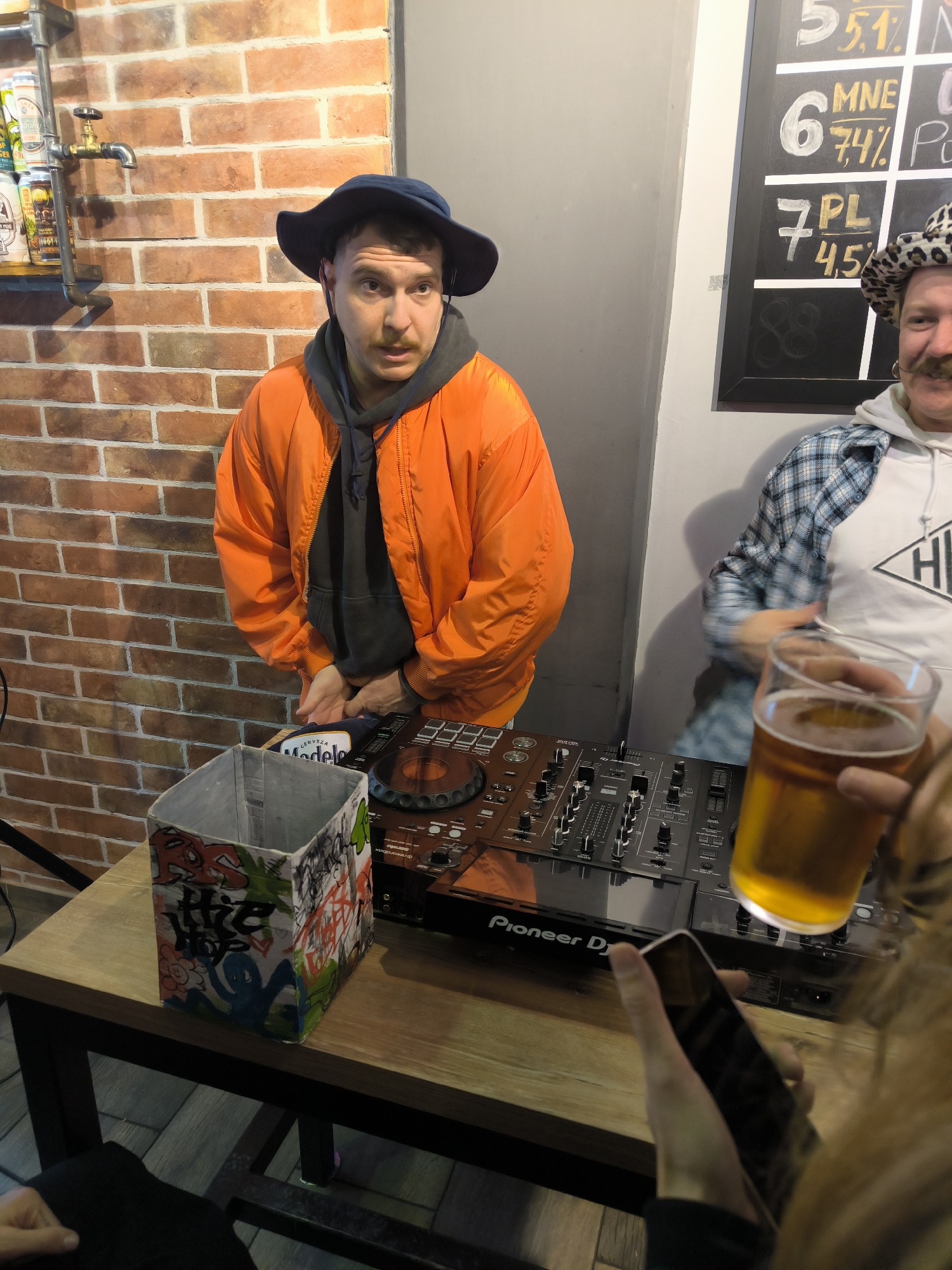 The scene depicts an indoor bar environment in Bar, Montenegro, during what appears to be a DJ performance.
In the foreground, a person's hands are visible on the right, holding a clear pint glass filled with an amber liquid (likely beer) in the left hand and a dark smartphone with a dark screen in the right hand.
To the left of the foreground, on a light-colored wooden table with dark metal legs, a square-shaped container, possibly cardboard, is present. Its interior is light grey, while its exterior is covered in colorful graffiti tags and illustrations, including elements like "RS," "tHe Hop," and a blue octopus-like figure. Adjacent to this box, a black Pioneer DJ controller with numerous knobs, faders, and illuminated buttons is set up.
Behind the table, a male individual with a light mustache, wearing a dark blue bucket hat, an orange bomber jacket, and a dark grey hooded sweatshirt, stands centered. His hands are clasped together above the DJ equipment, and he is looking slightly to the upper right with a focused or slightly surprised expression.
To the far right, a second male is partially visible. He has a light-colored mustache and is wearing a leopard print bucket hat, a light blue and white plaid shirt over a white hoodie with a black geometric logo (appearing as "HLD" or "HIE"). He is smiling and looking towards the left.
The background on the left features a reddish-brown exposed brick wall. A metal pipe assembly with a brass-colored spigot is affixed to this wall above a shelf holding several beer cans, one of which displays "Cerveza Modelo." The wall transitions to a smooth light grey surface on the right, against which a black chalkboard menu is mounted. The chalkboard displays items written in white chalk, including "5 5,1%", "6 MNE 7,4%", "7 PL 4,5%", and an "8" with an unreadable symbol below it, likely indicating drink options and alcohol percentages. The lighting is warm and artificial.