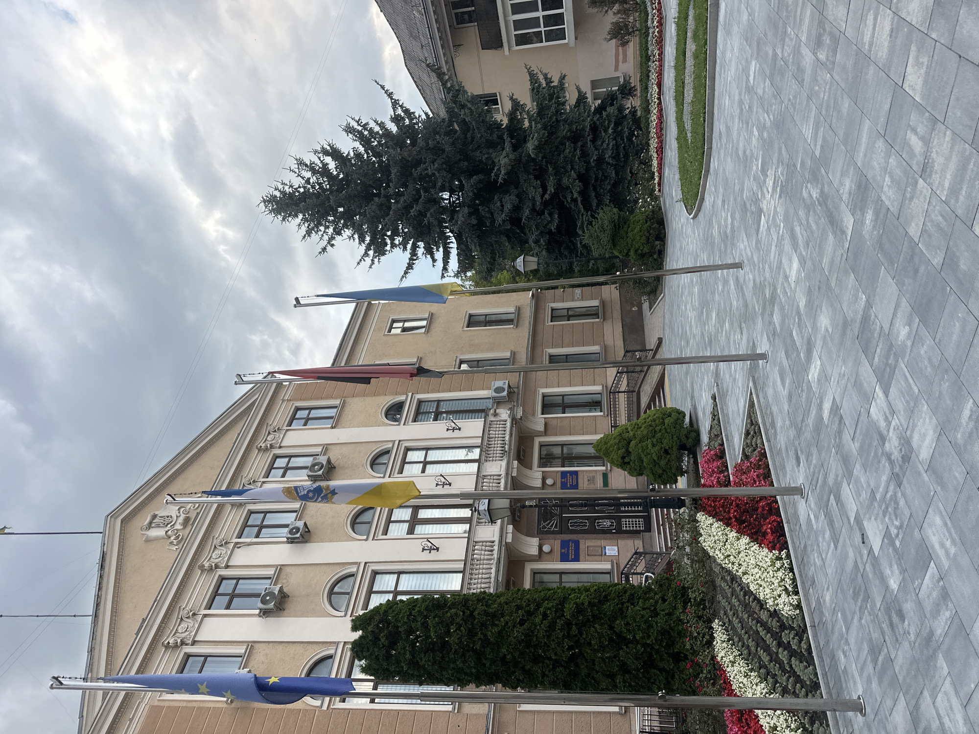 The image shows the Ternopil City Council building, located at 1 Teatralna Square, Ternopil, Ukraine. The building is a multi-story structure with a beige facade, featuring arched windows and decorative elements. In front of the building, there is a paved courtyard with several flagpoles displaying various flags, including the European Union flag, the Ukrainian flag, and other unidentified flags. A large evergreen tree stands to the right of the building, and a flowerbed with red, white, and pink flowers is visible in the foreground. The sky is overcast with clouds. There are no people visible in the image.