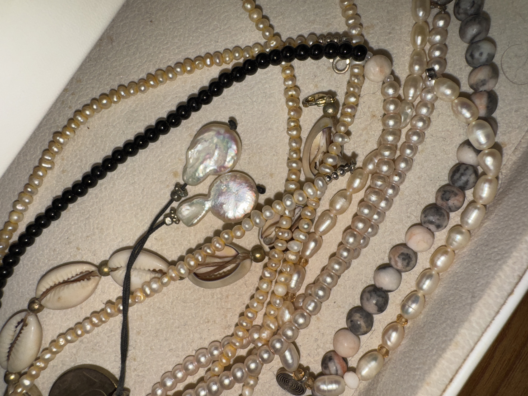 A collection of necklaces is displayed against a light-colored, slightly textured background, possibly fabric or a padded surface. The necklaces are diverse in style and materials, including strands of small, uniform pearls, black beads, cowrie shells interspersed with small pearls, irregularly shaped pearls, and larger stone beads with a mottled gray and pink coloration. One necklace features iridescent pearl pendants attached by thin black cords. Metal clasps and small metal components are visible on several of the necklaces. The overall arrangement is scattered, suggesting the items are being stored or displayed. There is no evidence to suggest a location in Kuta Selatan, Indonesia.