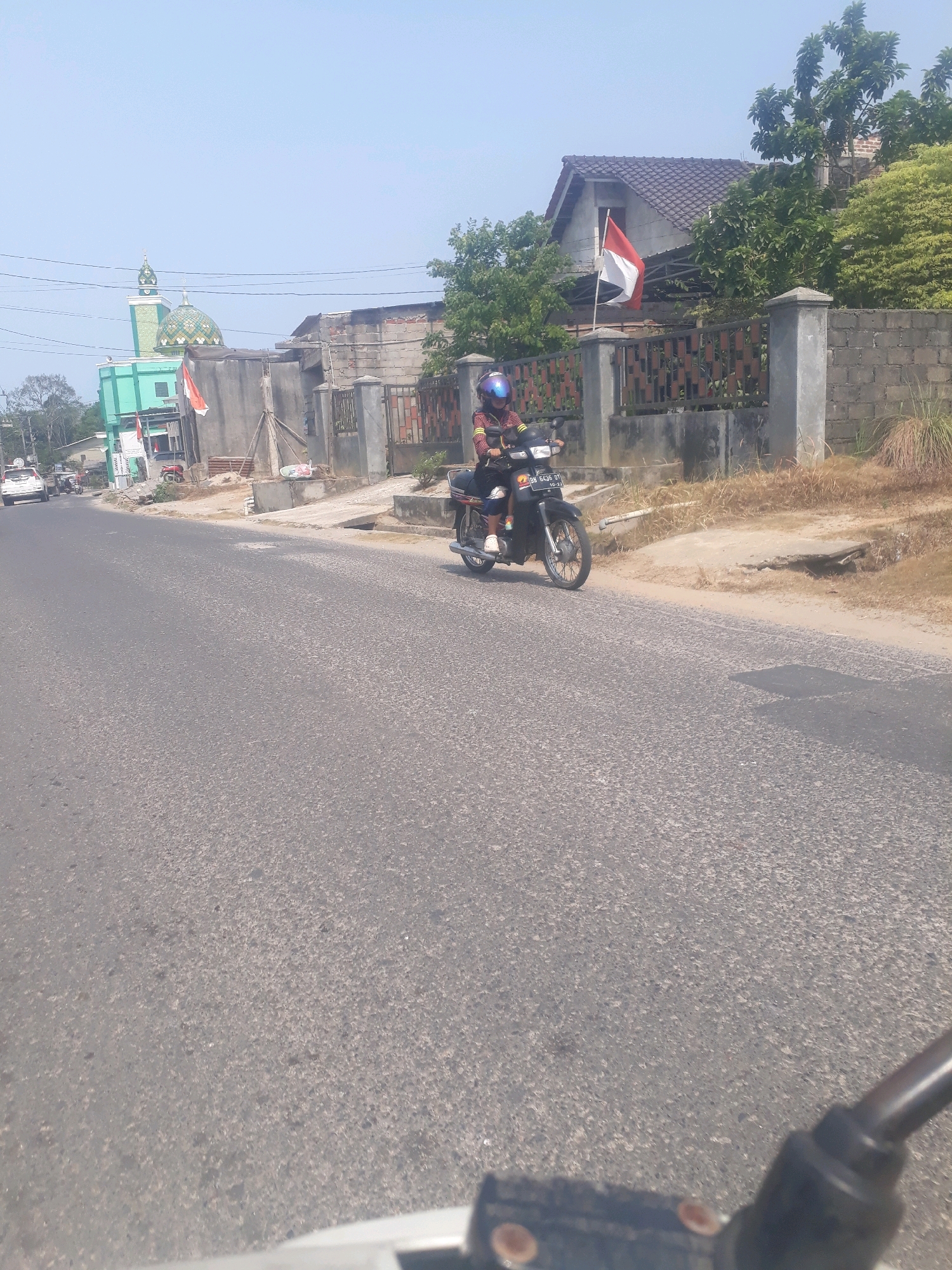 A person is riding a motorcycle down a paved road in Pangkalpinang, Indonesia. The rider is wearing a blue helmet and a yellow and red striped shirt. They are on a black motorcycle. In the background, there is a mosque with a green dome and minaret. There are also houses, fences, trees, and other buildings. The sky is clear and blue. The road appears to be in good condition. There is a flag on a pole in front of one of the houses. The motorcycle has a license plate that reads "BN 6436 GT". The scene appears to be a typical street scene in a city in Indonesia.