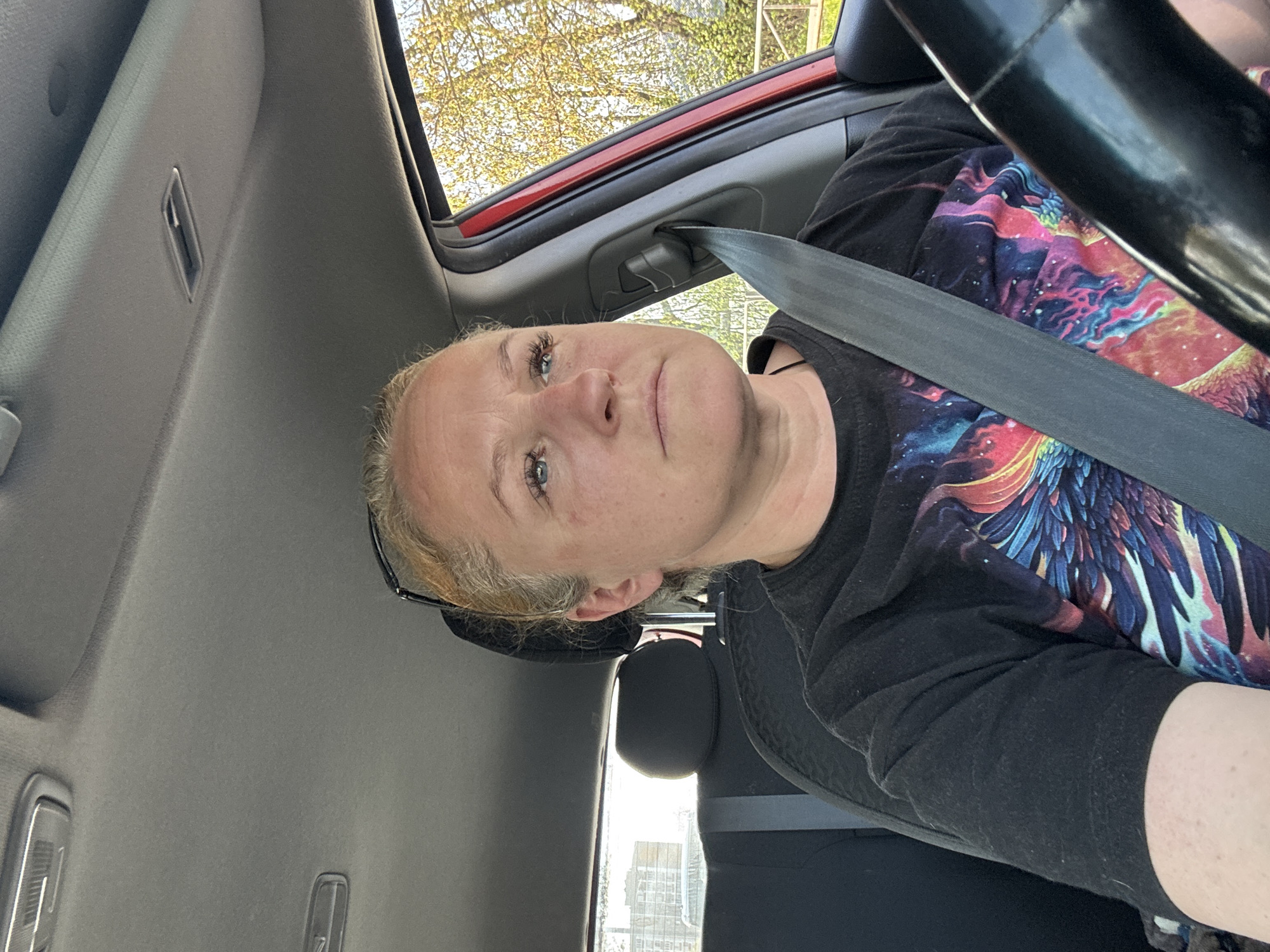 A woman is sitting in the driver's seat of a car, wearing a seatbelt and a black t-shirt with a colorful design. The car's interior is visible, including the steering wheel, roof, and a red window frame. Outside the car, trees are visible. There is no information in the image to indicate that the location is Chaikovsky, Russia.