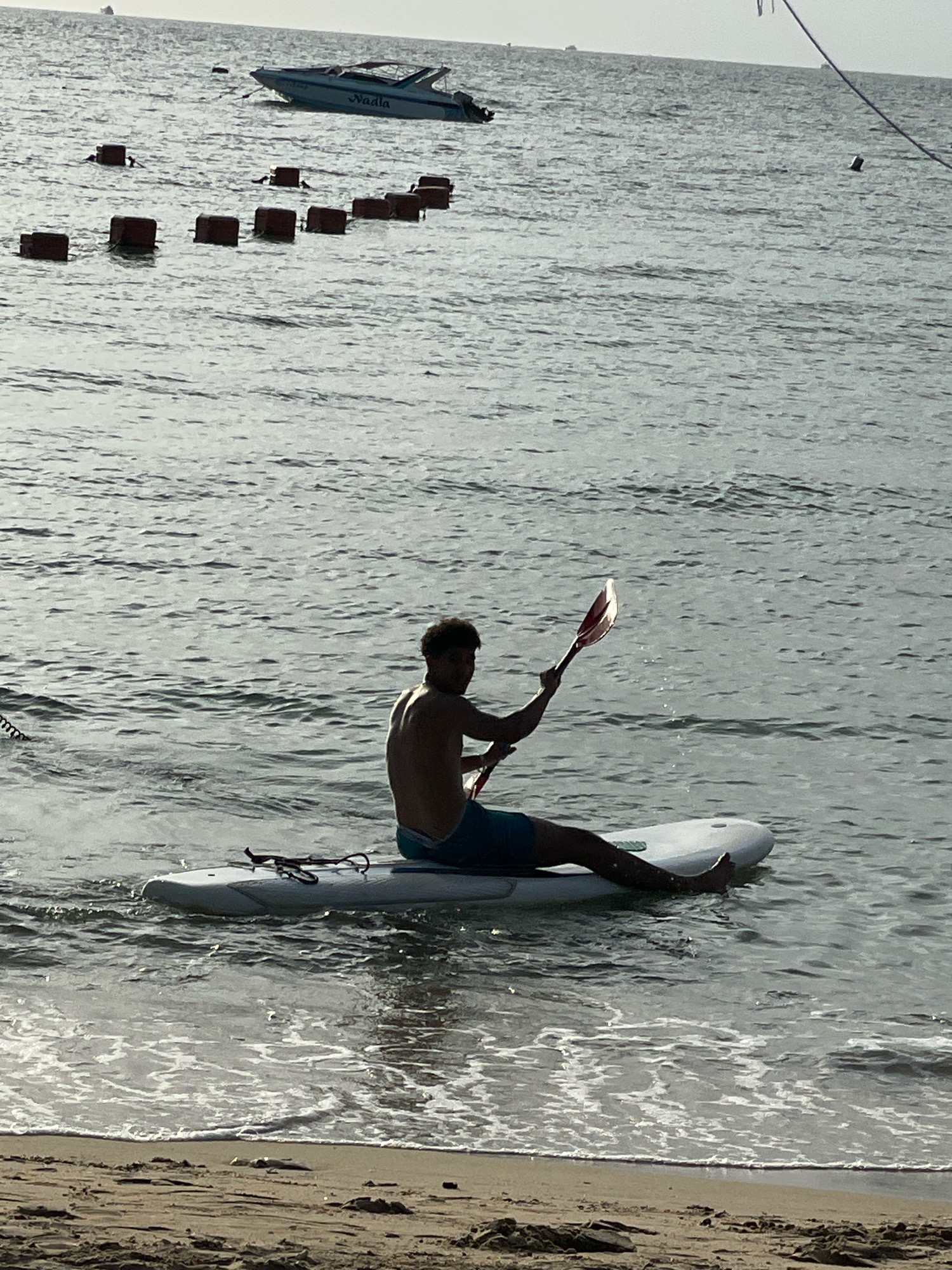A person sits on a paddleboard in the water near a sandy beach. The person, silhouetted by the sun, holds a paddle over their right shoulder. They wear blue shorts and appear to be in the process of paddling. The paddleboard is white and has a rope tied to it. The water is calm with small ripples, and the sandy beach is visible in the foreground with some debris. In the background, a white speedboat with "Naula" written on its side is anchored, and a series of red buoys are floating in a line. The sky is overcast. The scene is likely at Jomtien Beach in Pattaya City, Thailand, a popular spot for water activities.
