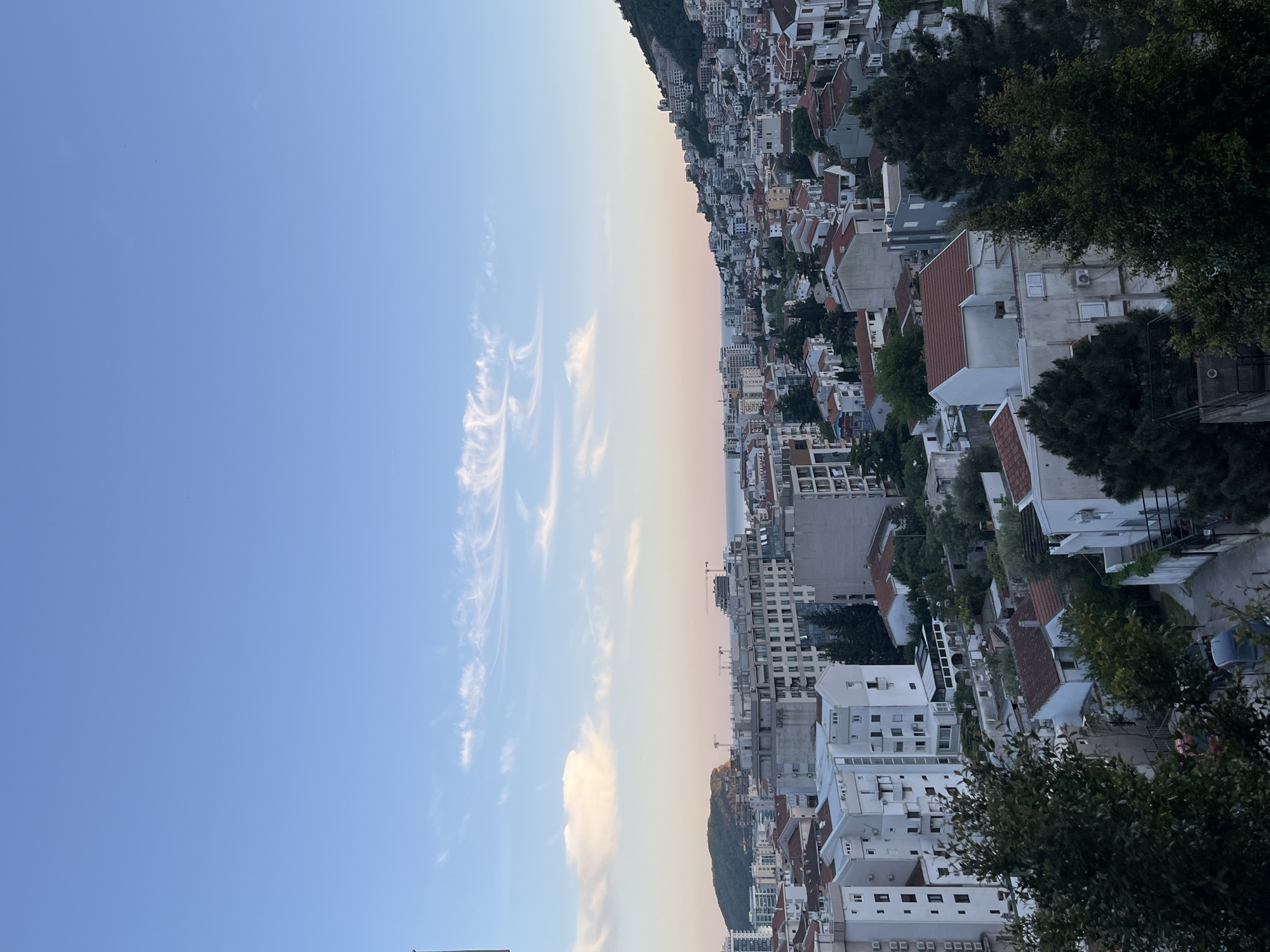 This media file captures a panoramic view of a coastal city, likely Budva, Montenegro, nestled against a mountainous backdrop under a vast sky during either sunrise or sunset.
The upper portion of the image is dominated by the sky, transitioning horizontally from a clear, vibrant blue on the left to softer, warmer hues of light pink and orange towards the right horizon, indicative of a low sun angle. Wispy, white cirrus clouds are scattered across the bluer section of the sky, while a larger, fluffy white cloud is visible closer to the warmer horizon.
Below the sky, the city sprawls across the frame, with dense clusters of buildings climbing up the slopes of a verdant mountain. The architecture predominantly features light-colored, often white, buildings with distinctive red-tiled roofs. Interspersed among these are some taller, more modern multi-story structures, with a few construction cranes visible, suggesting ongoing development. Lush green trees are dotted throughout the cityscape and cover the mountain slopes. A narrow strip of the calm sea is visible in the distance along the horizon line.
No specific activity or event is taking place, and no people are visibly identifiable. The overall impression is one of a peaceful, expansive landscape view. There is no visible text in the image.