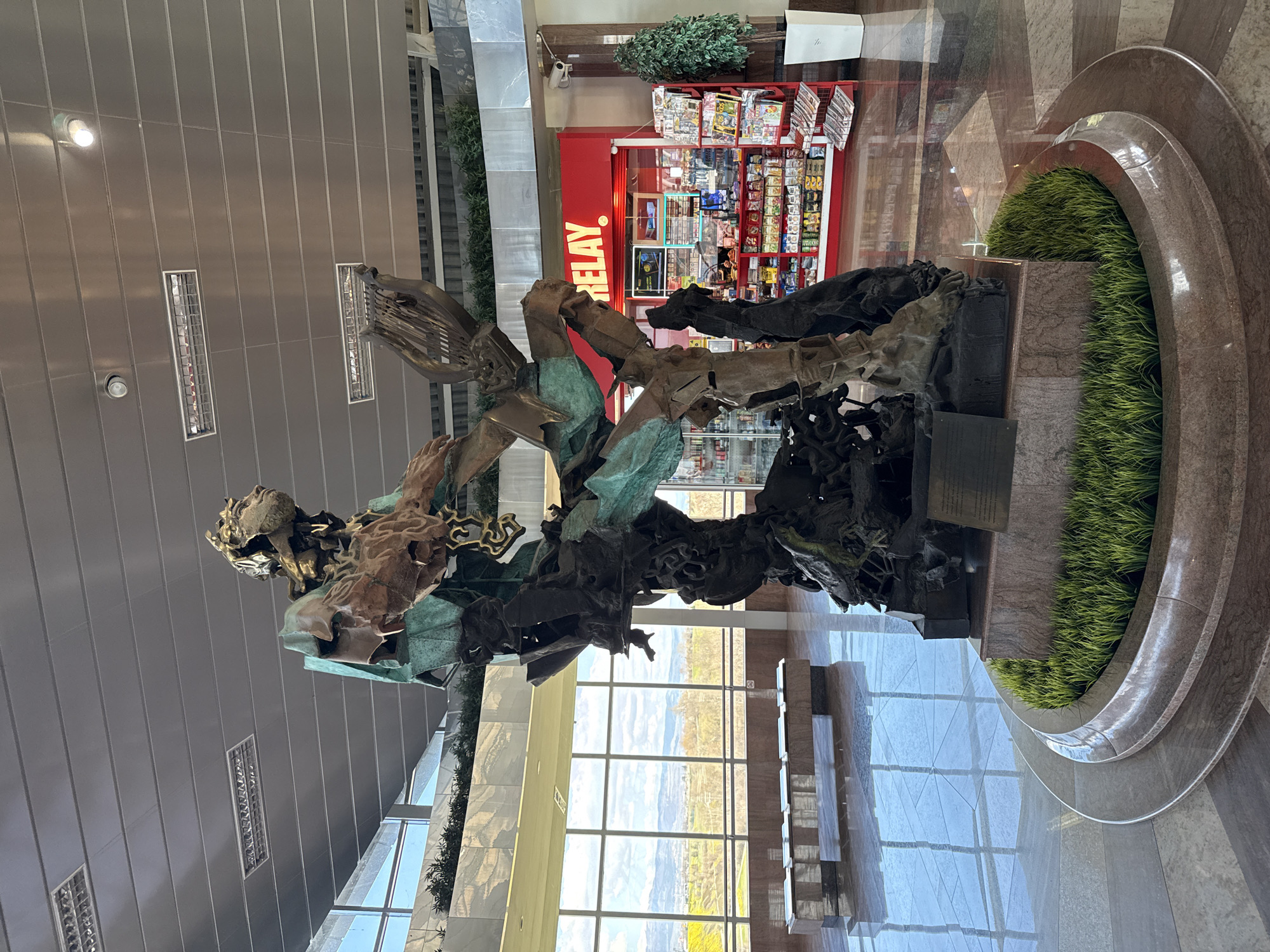 The image shows a large metal sculpture in an indoor space with large windows. The sculpture features human-like figures, with visible details like hands and faces, but appearing abstract and fragmented. The sculpture is set in a circular planter with greenery. Behind the sculpture is a 'Relay' branded store selling newspapers, magazines and other goods. Outside the windows there appears to be a landscape with fields and hills.
The scene is likely in an airport building in Sofia, Bulgaria.