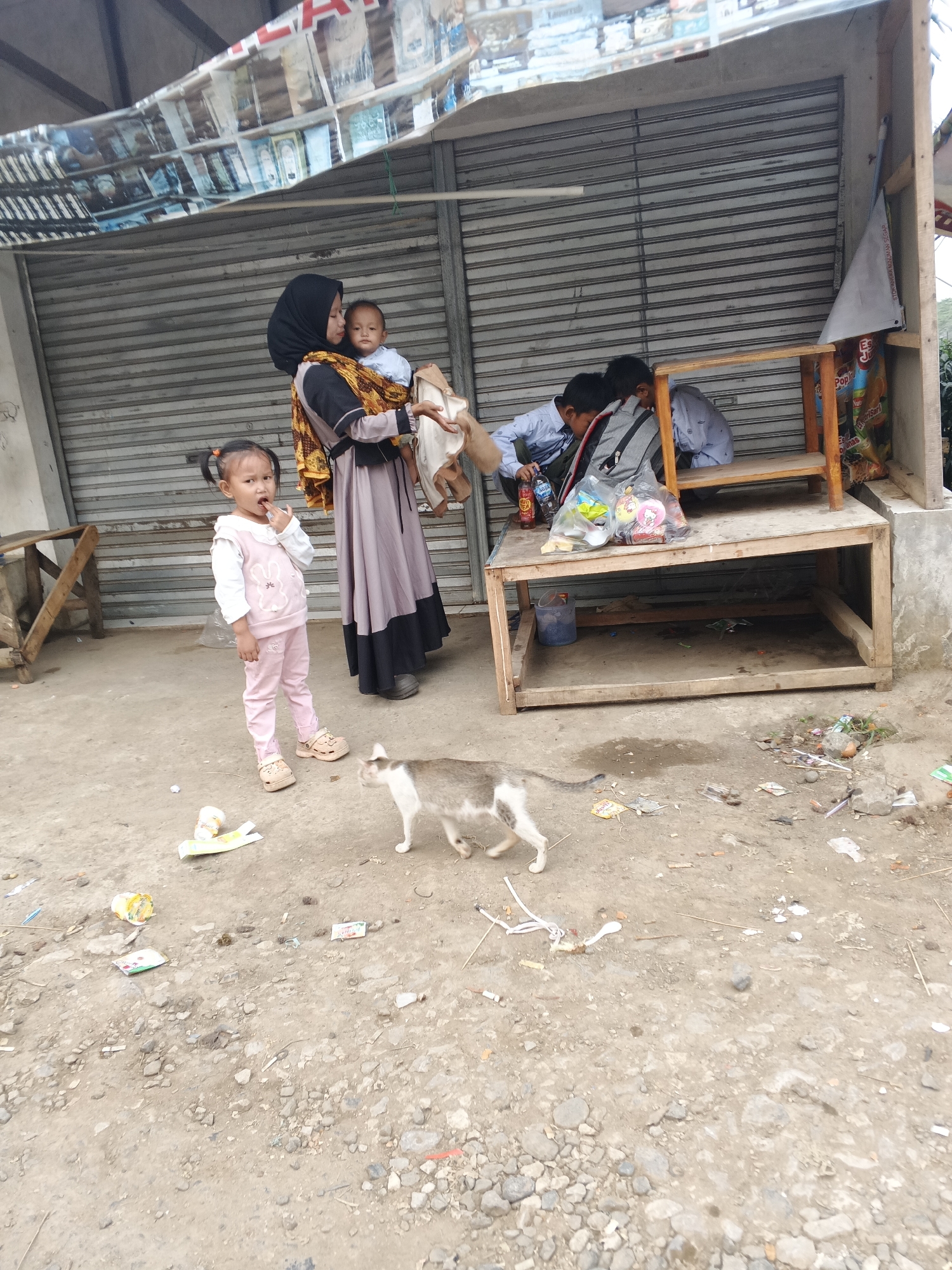 The image depicts a scene outside a storefront in Warungkiara, Indonesia. A woman wearing a hijab and a long dress is holding a baby. To her left, a young girl in pink pants and a pink vest stands with her finger in her mouth. In the background, two young boys are seated at a wooden table. The ground is dirty and scattered with debris. A cat walks across the ground in the foreground. The storefront has a metal shutter and a wooden structure with shelves. Above the shutter, a banner with text and images is displayed.
