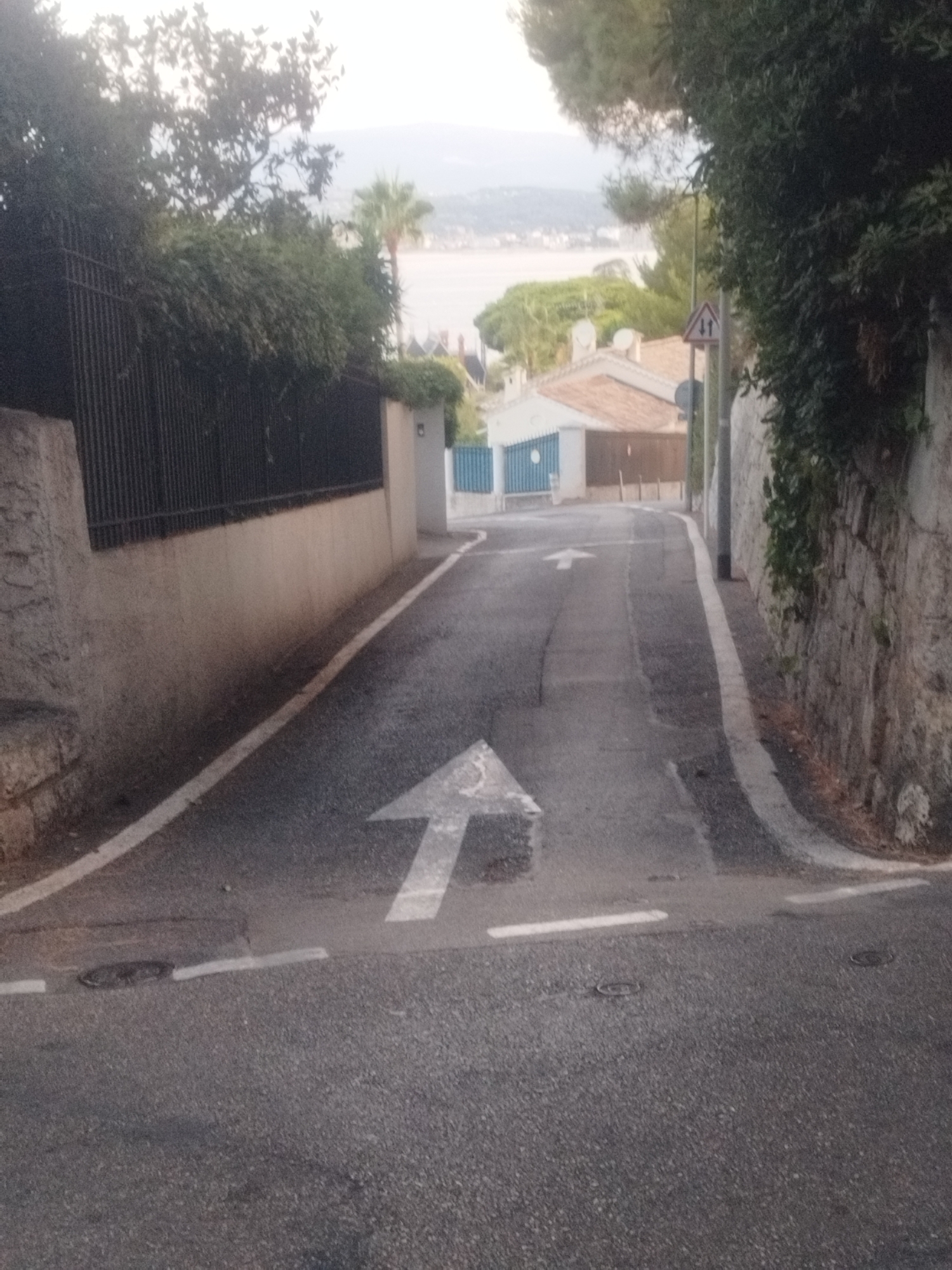 The image depicts a street in Antibes, France, as evidenced by a street sign featuring a symbol commonly used in France and the presence of Mediterranean flora. The scene shows a sloped asphalt road with white lane markings, including a prominent white arrow pointing upwards, indicating the direction of travel. The road is bordered by a stone wall on the right and a metal fence with vegetation on the left. In the background, buildings with terracotta roofs and a body of water are visible, suggesting a coastal location. The overall impression is that of a quiet, residential street.