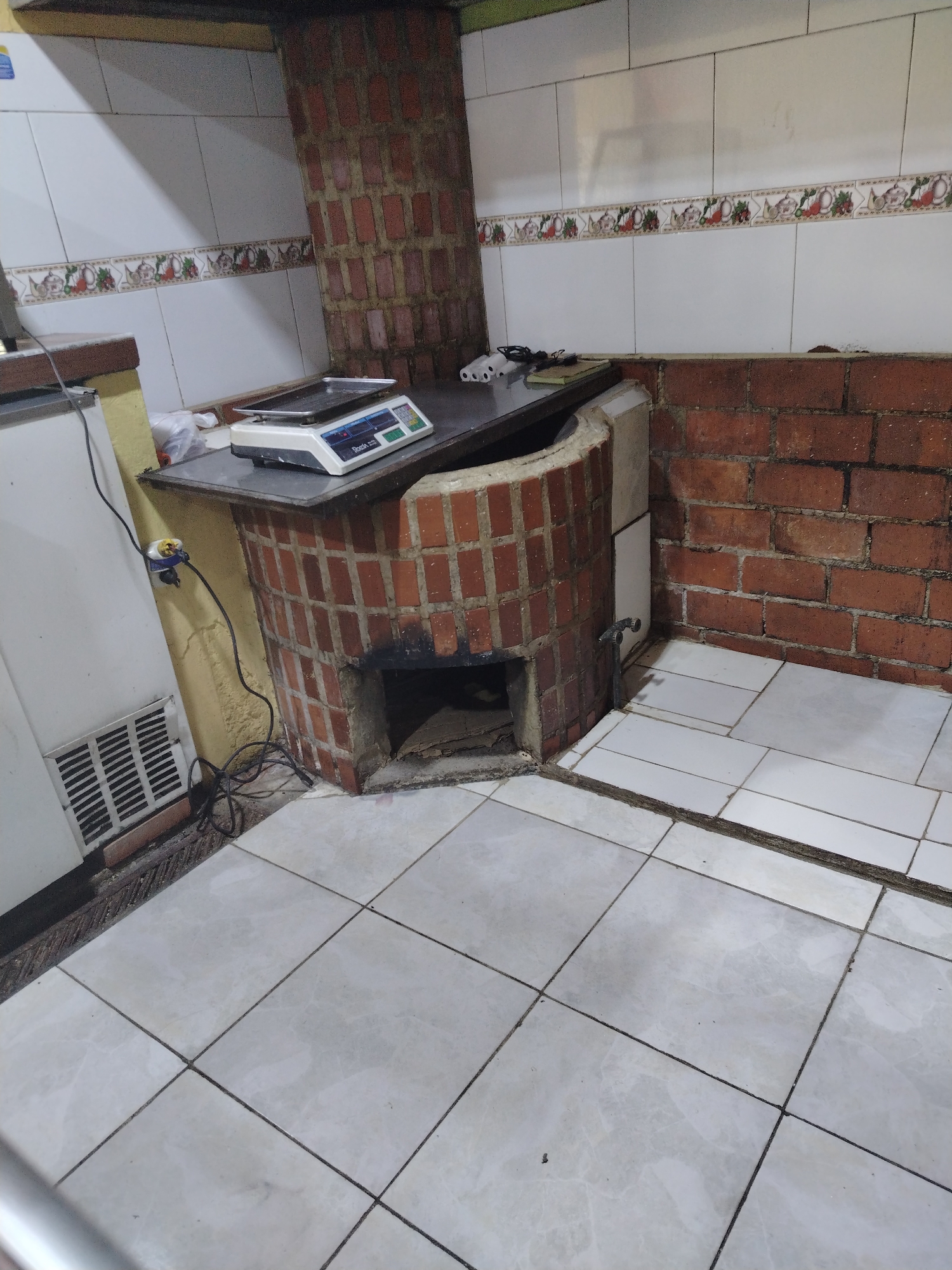 This image captures an indoor scene, likely a kitchen or food preparation area within a small business in Tinaquillo, Venezuela. The setting features white tiled walls with a decorative border showcasing illustrations of fruits and teapots. The floor is covered in light-colored ceramic tiles, which appear worn and somewhat dirty, with dark grout lines.
A dominant feature is a rustic, brick-built structure, possibly a traditional wood-fired oven or grill, with a chimney extending upwards. At its base, a dark opening serves as a firebox or ash pit, containing some debris. A small metal pipe or faucet is affixed to the side of this brick structure.
On a counter surface integrated into the brickwork, a white digital weighing scale with a silver platform is placed. The brand name "Boada" is visible on the scale's display area. Nearby, a small dark rectangular object and a white plastic bag can be seen. To the left, a white refrigerator or freezer is positioned, featuring a ventilation grille at its lower section. An electrical outlet with a yellow plug is visible on the wall, with tangled black wires accumulating on the floor. No individuals are present in the image, but the arrangement of objects suggests a space actively used for cooking or commerce. The overall impression is one of a functional, utilitarian space that appears a bit disheveled.