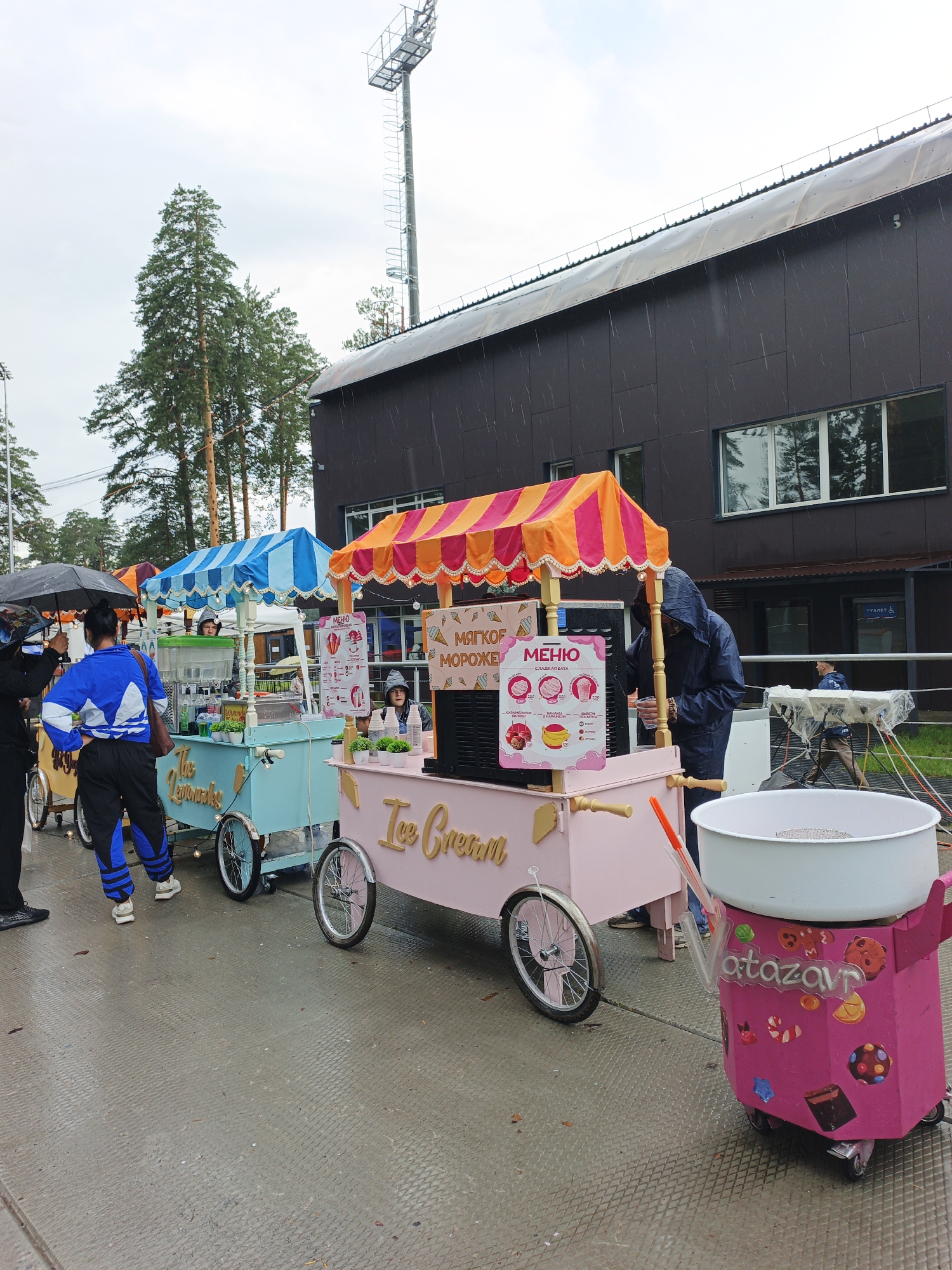 The image depicts an outdoor scene in Zlatoust, Russia, on a rainy day. The foreground features several brightly colored ice cream carts lined up. The cart on the right is pink and decorated with candy stickers and the word "atazavr." It has a large white bowl on top, likely for cotton candy. To its left is another pink ice cream cart with "Ice Cream" written in cursive on its side. Further left is a light blue ice cream cart. Each cart has a colorful awning.
Several people are present. One person in a blue tracksuit is standing with their back to the camera, near the blue cart. Another person in a dark hooded raincoat is standing at the pink cart, appearing to be serving a customer. The menu board on this cart displays various ice cream options. A person holding a black umbrella is visible on the far left. In the background, a large, dark-colored building with a distinctive curved roof can be seen, along with tall pine trees. A stadium light tower is also visible against the overcast sky. The ground appears to be paved and wet, indicating the rain. The overall atmosphere suggests a fair or outdoor event taking place despite the weather.