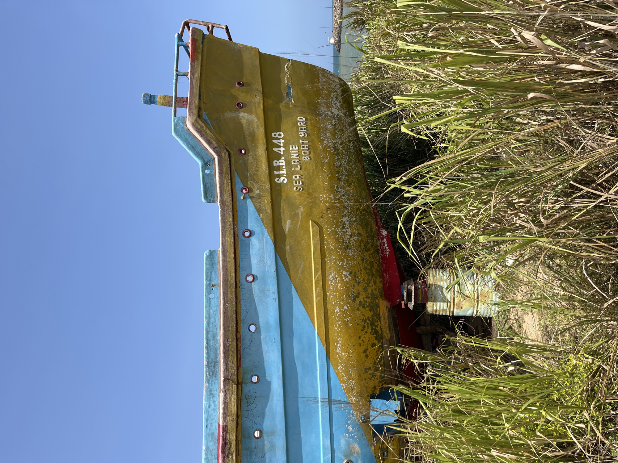 The image shows the stern of a disused boat, painted in bright blue and yellow, resting on dry land amongst tall, dry grasses. The boat has text written on its yellow hull that reads "S.L.B. 448 SEA LANIE BOAT YARD". The boat is supported by a rusty metal structure. In the background, the sea is visible, along with a small lighthouse or navigational marker. The sky is clear and blue. The scene appears to be an outdoor boatyard or storage area in Dondra, Sri Lanka, likely near the coast, given the presence of the sea and the boat. No people are visible in the image.